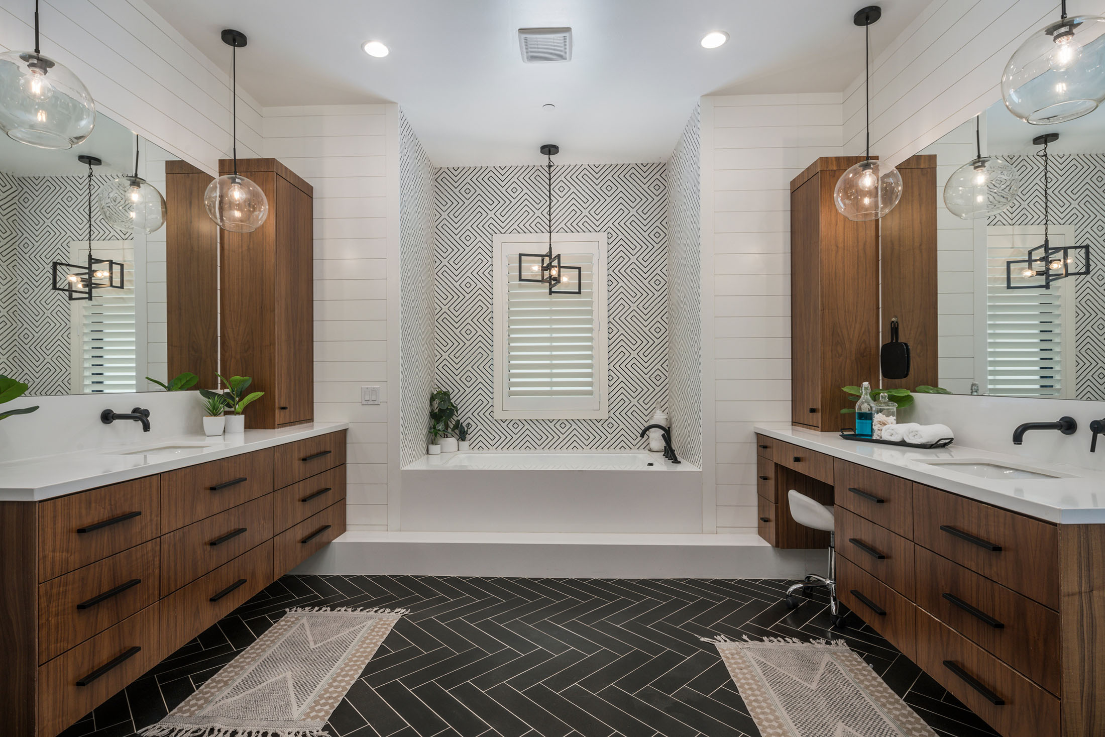 Modern Double Vanity Bathroom with Geometric Tile Design – A stylish bathroom featuring dual wooden vanities, geometric patterned backsplash, pendant glass lighting, and a built-in bathtub. The black herringbone tile flooring adds contrast, creating a luxurious and contemporary atmosphere.