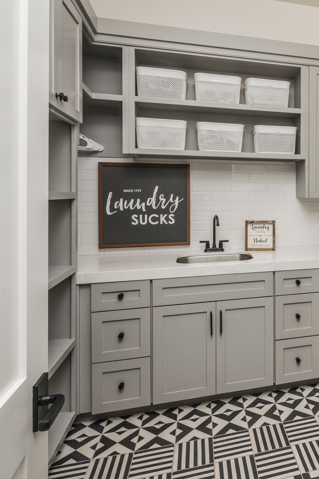 Modern Gray Laundry Room with Custom Cabinetry and Patterned Tile Floor – A stylish laundry room featuring gray shaker cabinets, open shelving with white storage bins, a farmhouse sink, and a black and white geometric tile floor. A framed sign humorously states "Laundry Sucks."