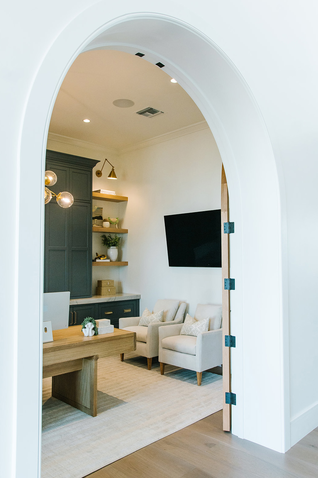 Sophisticated Home Office with Arched Doorway - A stylish home office with a natural wood desk, plush white armchairs, dark cabinetry, and floating shelves, framed by a striking arched entryway.