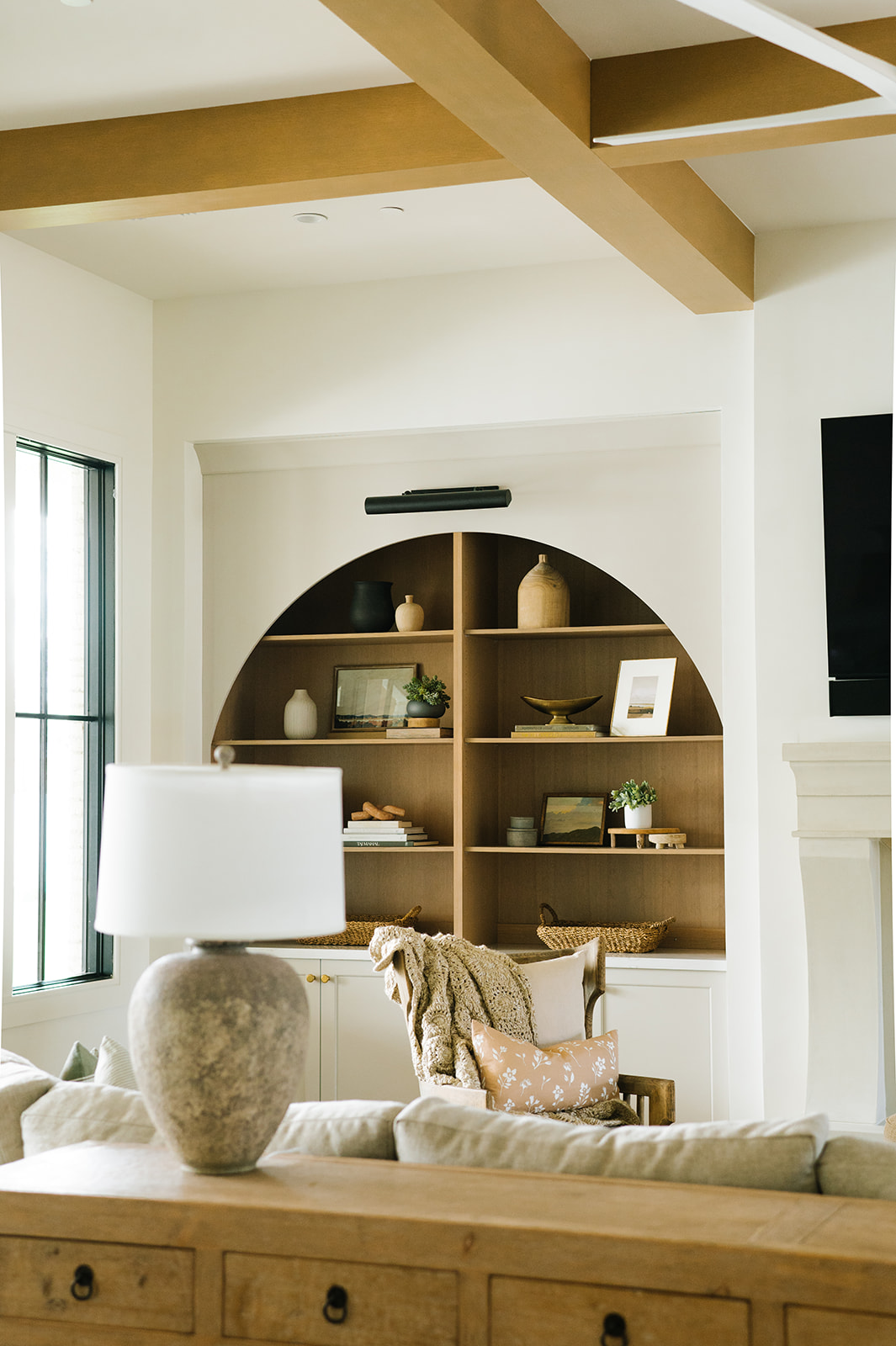 Elegant living space featuring a custom-built arched wood bookcase with warm tones, styled with curated decor and natural elements. The room showcases exposed ceiling beams, a cozy neutral-toned sofa, and layered textures, blending rustic charm with modern sophistication for a timeless aesthetic.