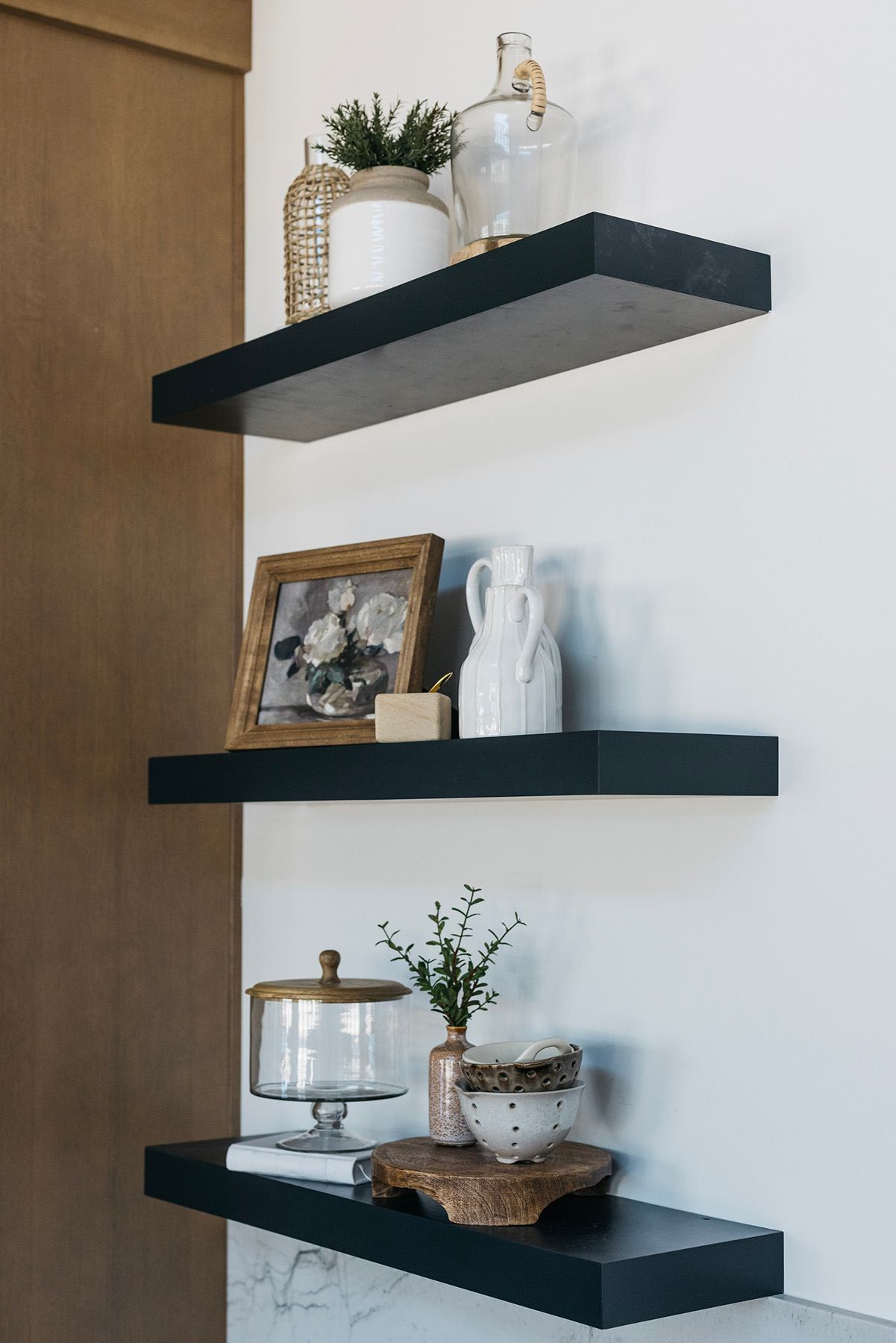 Modern Floating Shelves with Decorative Styling – Elegant black floating shelves adorned with neutral-toned ceramics, framed artwork, and greenery, enhancing this luxury kitchen or bathroom with curated home décor and stylish organization.