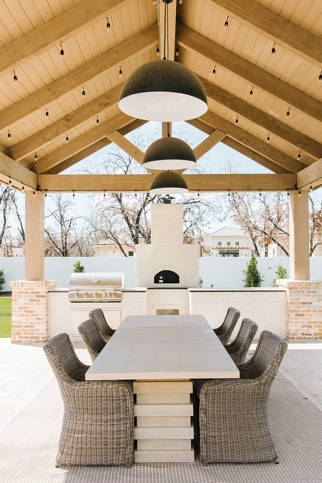 Outdoor dining area with a covered pergola, featuring a built-in stainless steel grill, white brick pizza oven, and a large wooden dining table with wicker chairs, illuminated by modern black pendant lights and string lights.