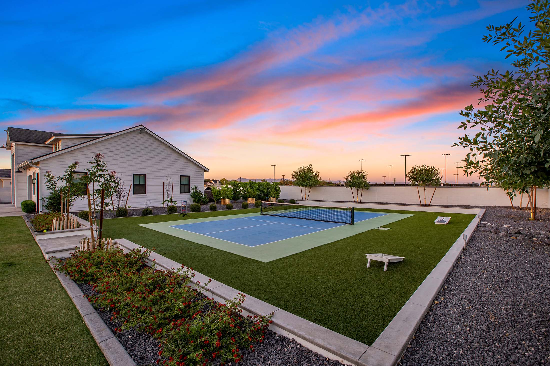 Expansive backyard featuring a private pickleball court, lush landscaping, and a modern farmhouse-style home. The serene outdoor space is designed for recreation and relaxation, with manicured greenery, vibrant flower beds, and stunning sunset views enhancing the luxurious setting.