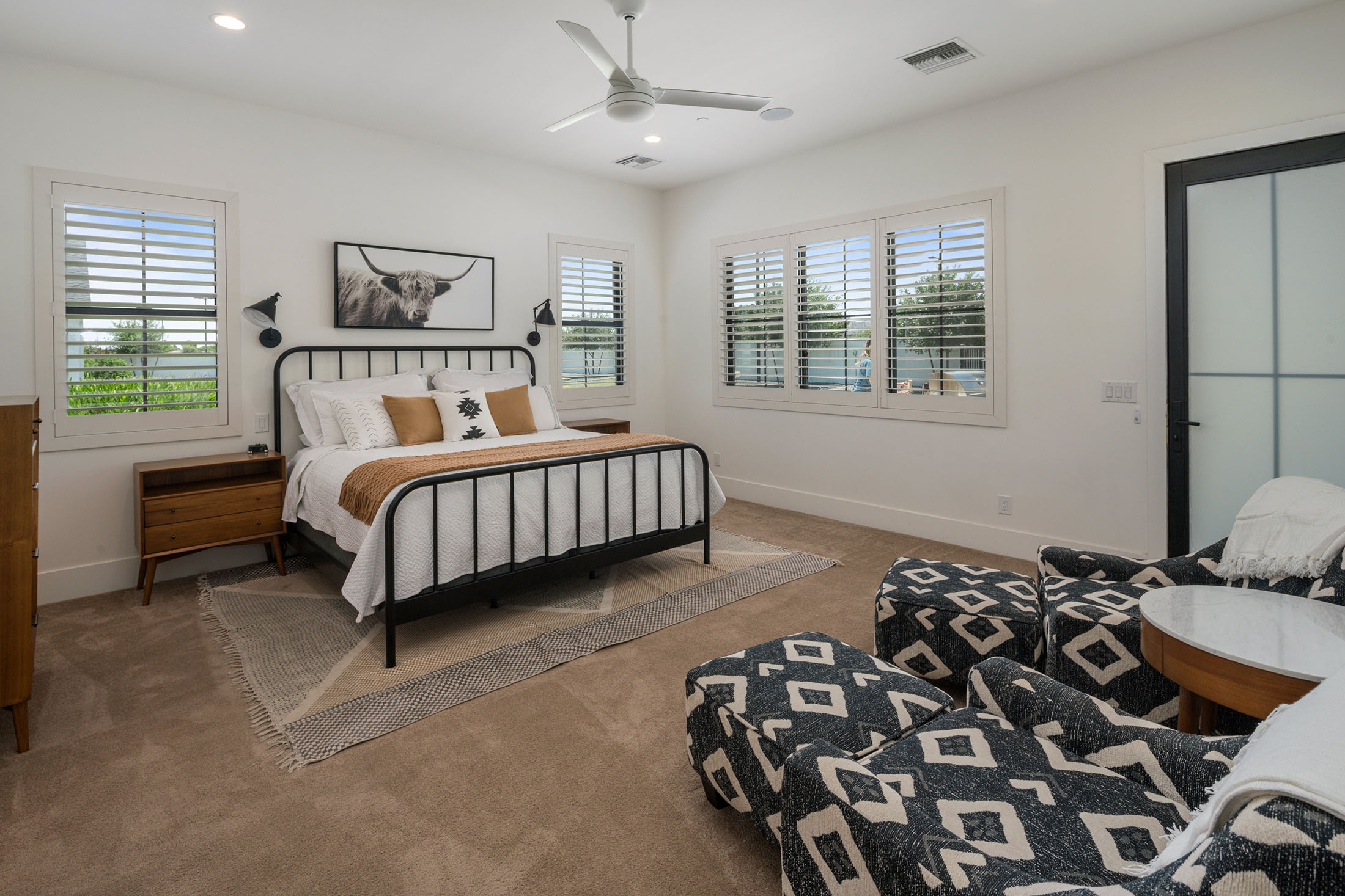 Minimalist Bedroom with Black Metal Bed and Southwestern Decor – A spacious bedroom with a modern black metal bed, neutral bedding with mustard accents, and a framed Highland cow print above the bed. Large windows with plantation shutters allow for abundant natural light.