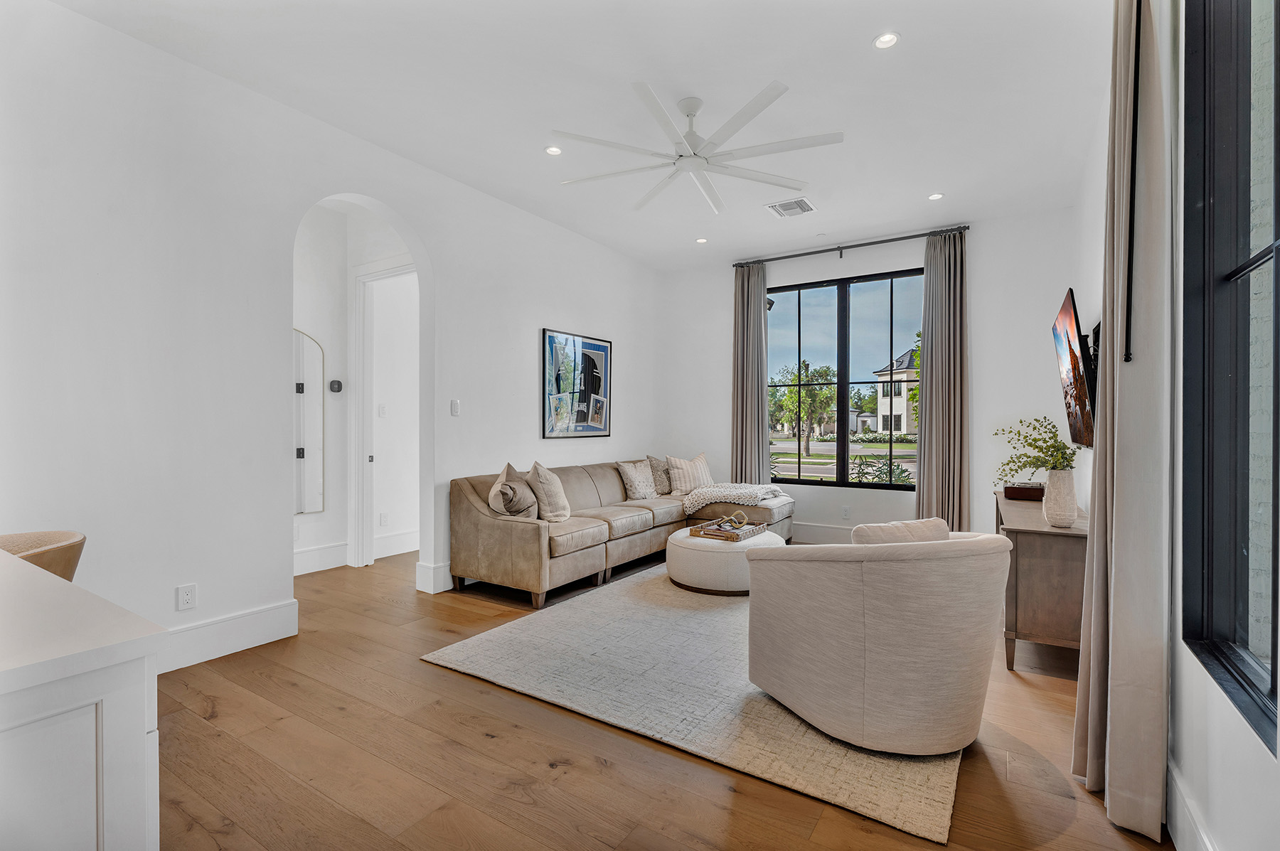 Bright and Elegant Living Room with Neutral Tones – A chic living space with a beige leather sofa, abstract wall art, and a round ottoman coffee table. Large windows with neutral drapes allow natural light to flood the room, enhancing its inviting ambiance.