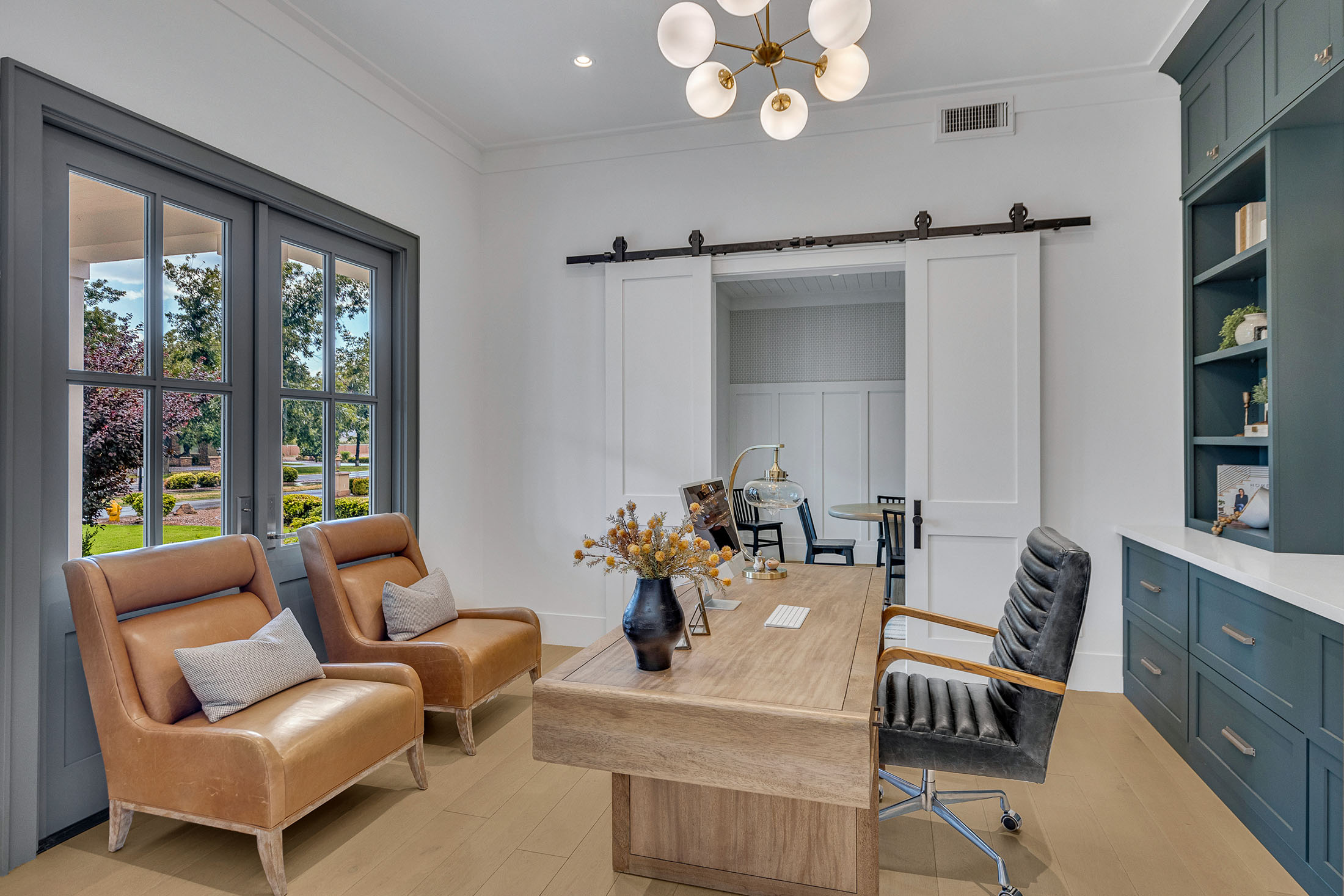 Sophisticated home office featuring a natural wood desk, built-in sage green cabinetry, and leather accent chairs. The space is illuminated by a modern brass chandelier and large glass-paneled double doors, while sliding barn doors lead to an adjoining room, blending functionality with timeless elegance.