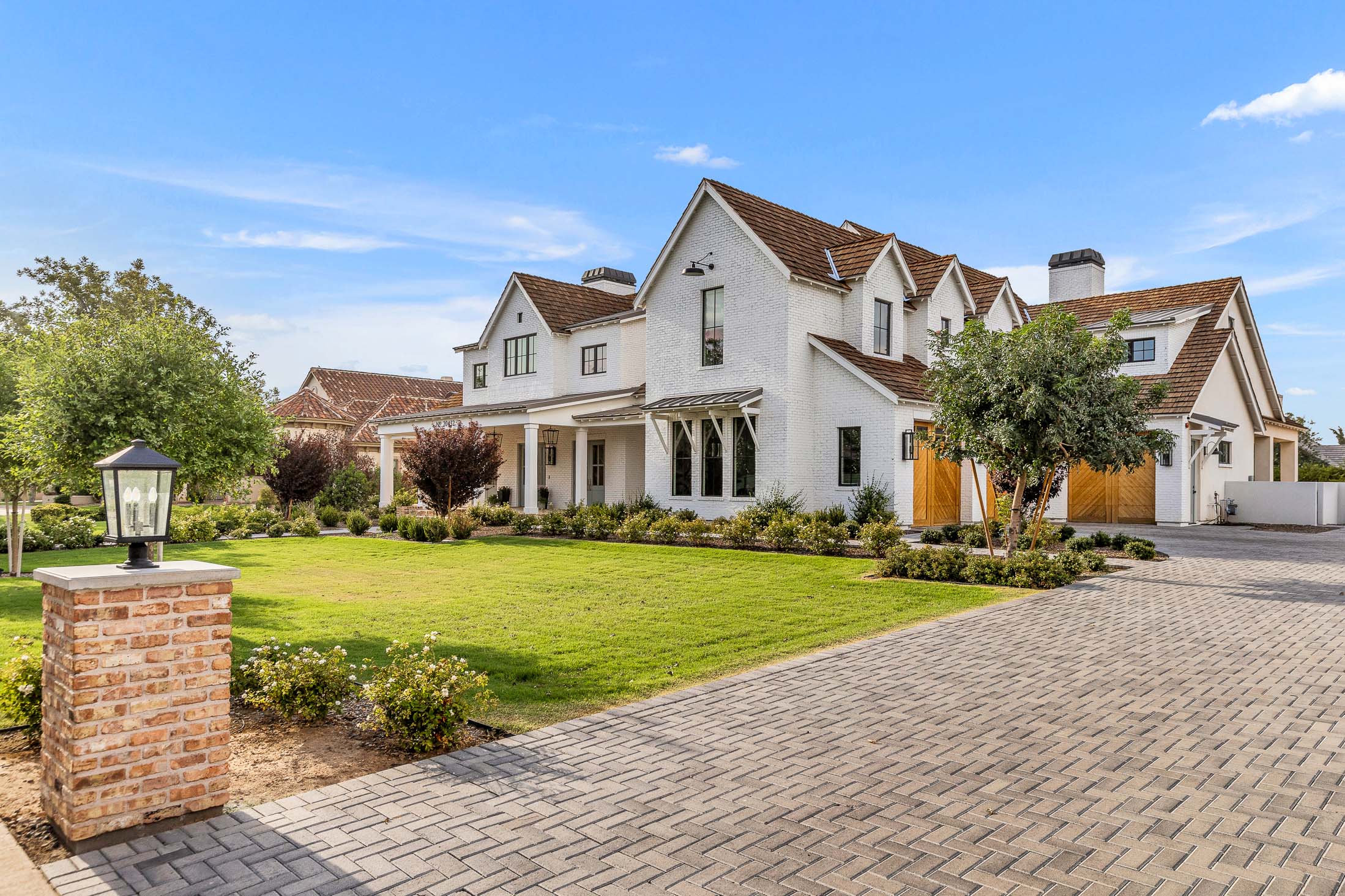 Luxury estate home with brick driveway and lush landscaping – A grand white brick home with a brown shingle roof, large windows, and wooden garage doors, complemented by a landscaped front yard and paver driveway.