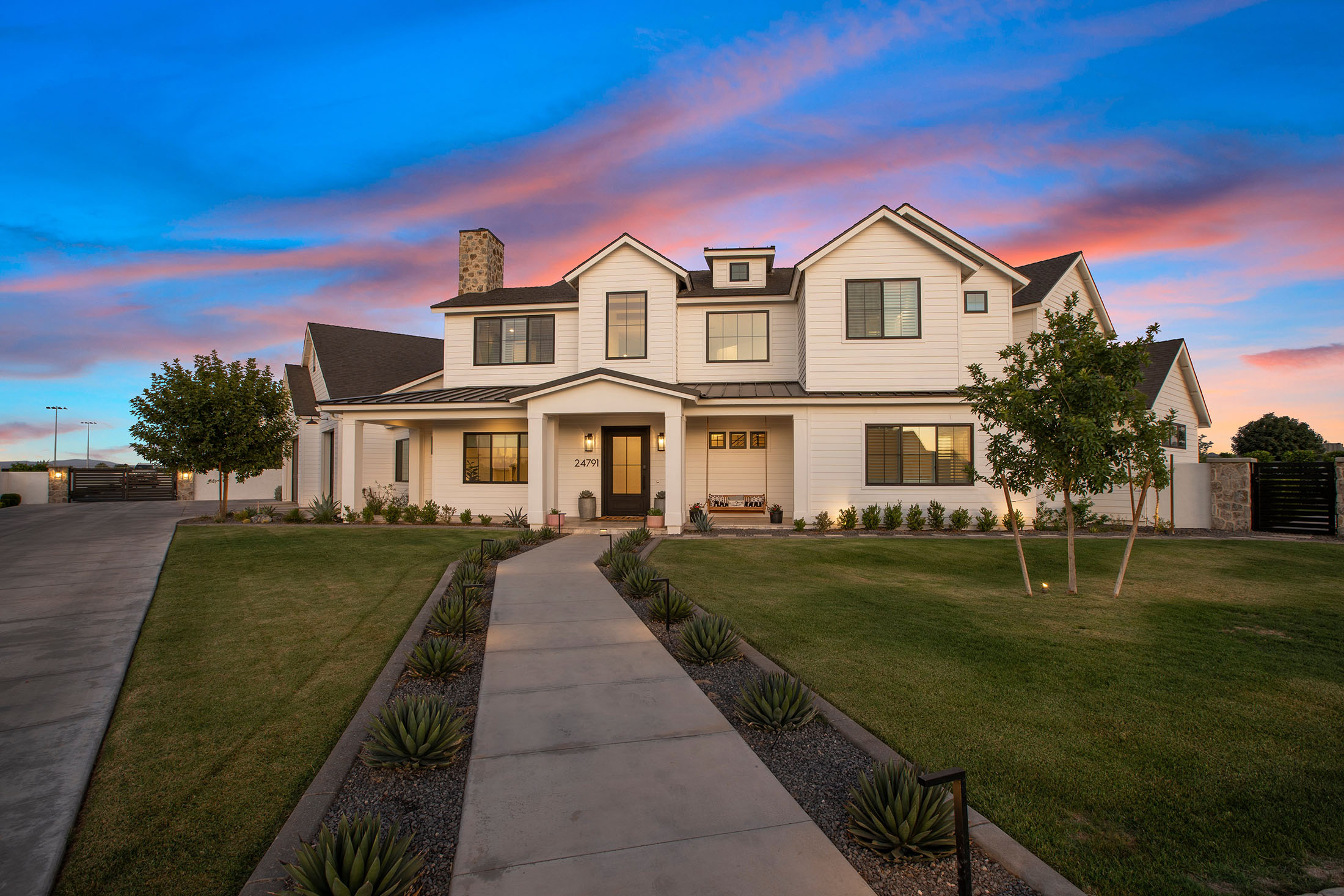 Modern farmhouse-style luxury home with a white board and batten exterior, black-framed windows, and a welcoming front porch. The beautifully landscaped front yard features a concrete walkway lined with agave plants, leading to the grand entrance. A stunning sunset sky enhances the home’s timeless elegance and curb appeal.