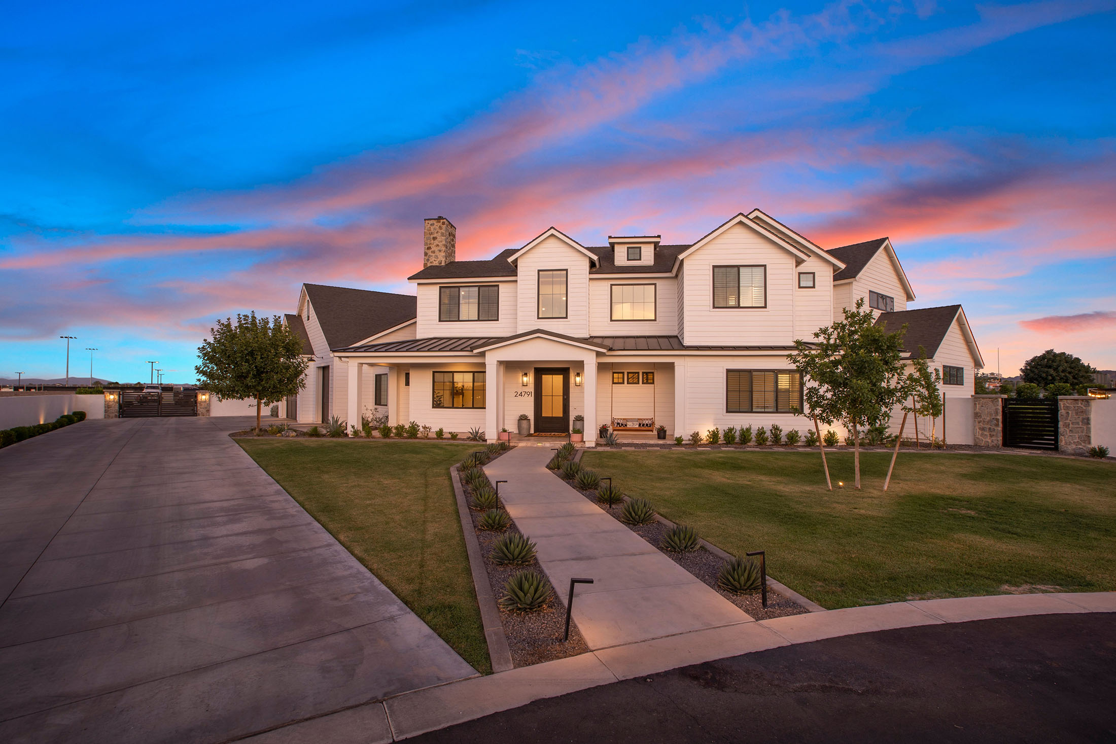 Stunning modern farmhouse exterior with sunset sky – A beautifully designed two-story white farmhouse with black trim, a metal roof, and a stone chimney, surrounded by lush landscaping and a long concrete driveway.