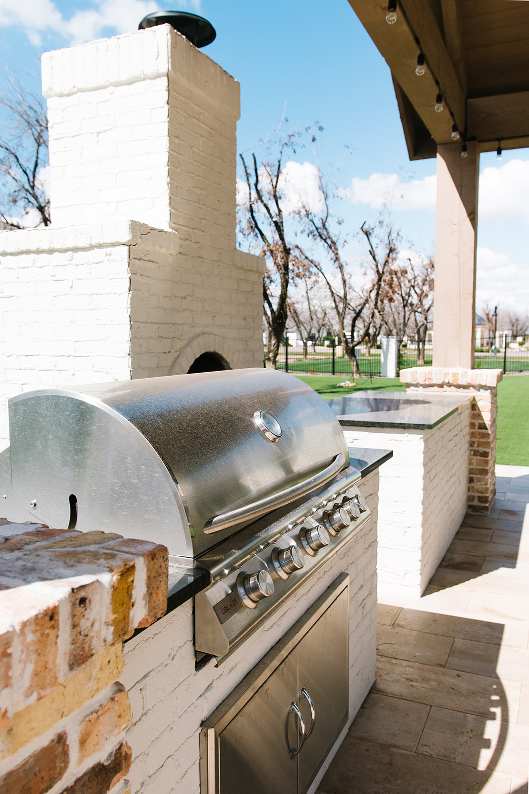 Outdoor Kitchen with Built-in Grill and Fireplace - A stylish outdoor kitchen featuring a built-in stainless steel grill, white brick fireplace, and a covered patio with string lights, set against a backdrop of lush greenery.