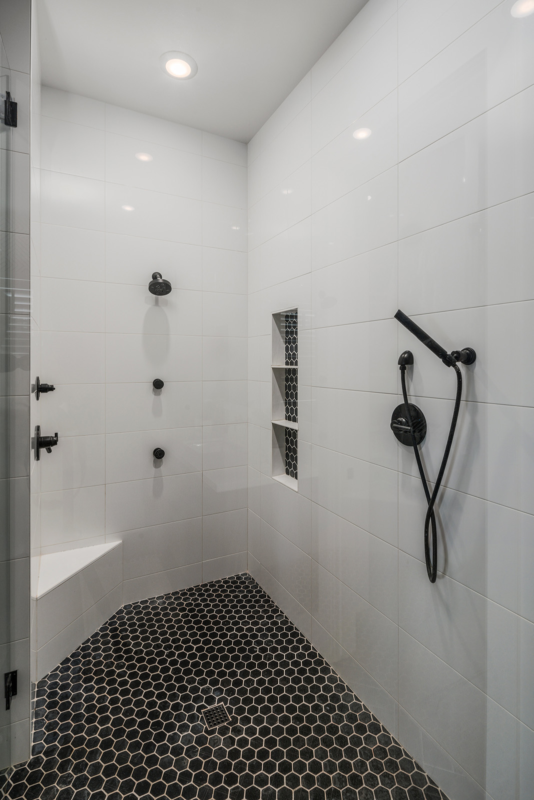 Minimalist Walk-in Shower with Black Hexagonal Tiles – A sleek, modern walk-in shower with glossy white tiled walls, black hexagonal tile flooring, and a built-in shower bench. The black fixtures add contrast, giving the space a sophisticated, minimalist appeal.