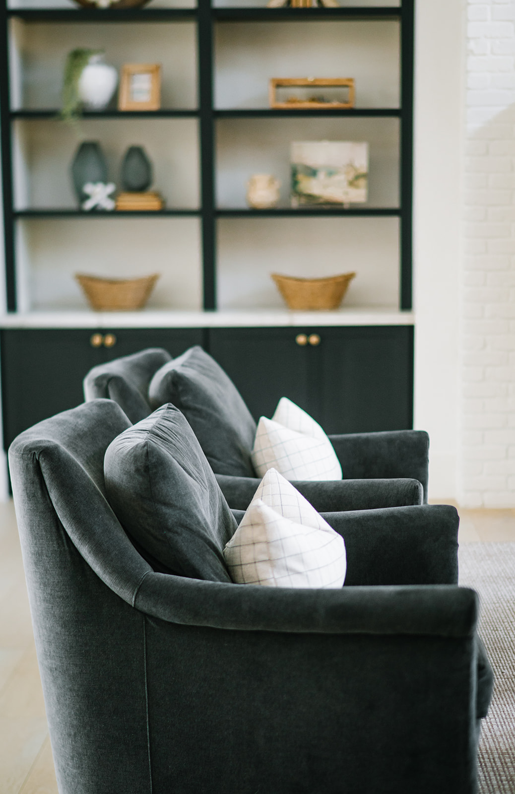 Sophisticated living space featuring plush charcoal velvet armchairs with patterned accent pillows, positioned in front of a striking black built-in bookcase with brass hardware and styled decorative elements. The combination of rich textures and contrasting tones creates a refined, modern aesthetic.