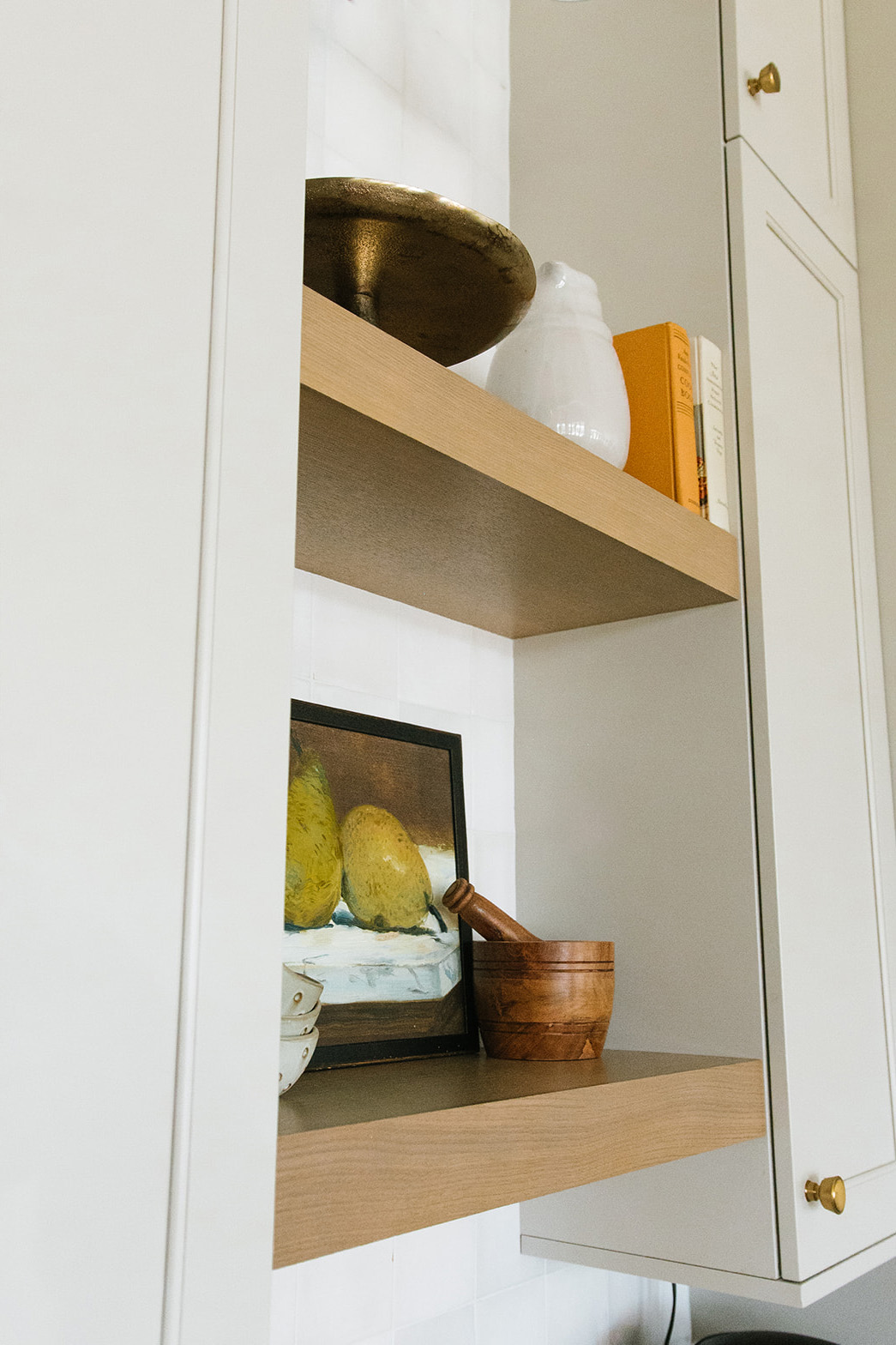 Floating Kitchen Shelves with Decorative Accents - A close-up of floating wood shelves in a modern kitchen, adorned with an art print of pears, a wooden mortar and pestle, ceramic vases, and neatly stacked books.