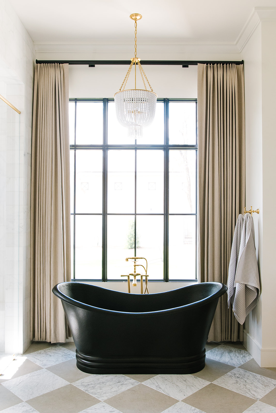 Elegant bathroom featuring a black freestanding soaking tub, a spacious glass-enclosed shower with gold fixtures, and floor-to-ceiling windows framed in black. The checkered marble flooring and crystal chandelier add a timeless, sophisticated touch.