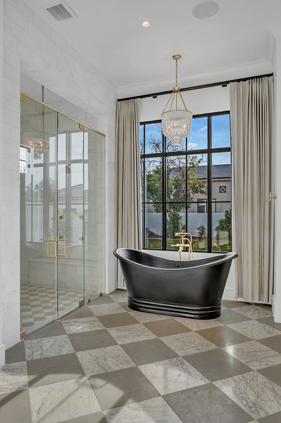Elegant bathroom featuring a black freestanding soaking tub, a spacious glass-enclosed shower with gold fixtures, and floor-to-ceiling windows framed in black. The checkered marble flooring and crystal chandelier add a timeless, sophisticated touch.