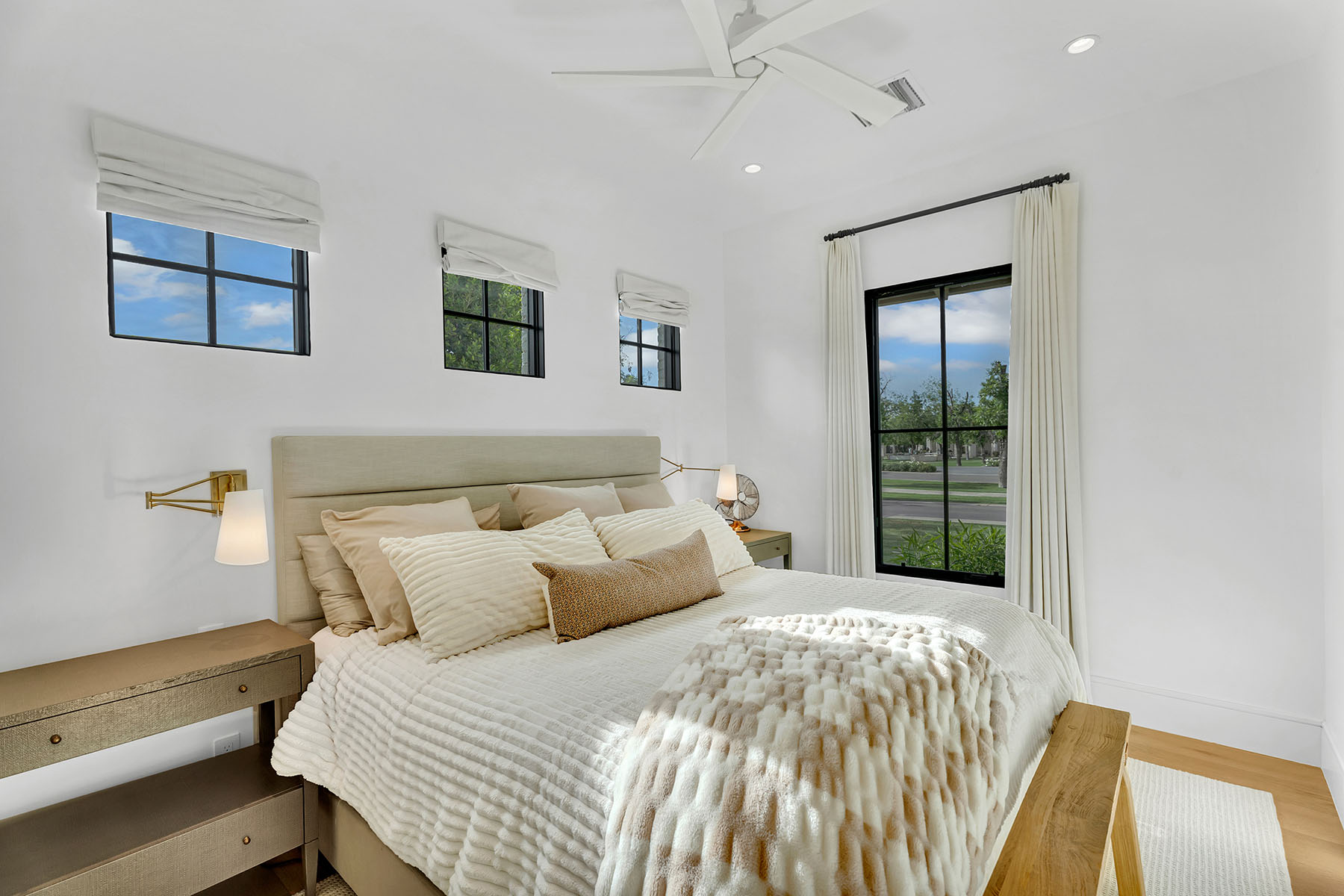 Cozy Neutral Bedroom with Soft Pink and Beige Accents – A beautifully styled bedroom featuring a light beige upholstered bed frame, soft pink and striped accent pillows, and brass swing-arm wall lamps. The modern bedside tables add a touch of elegance, complementing the minimalist decor.