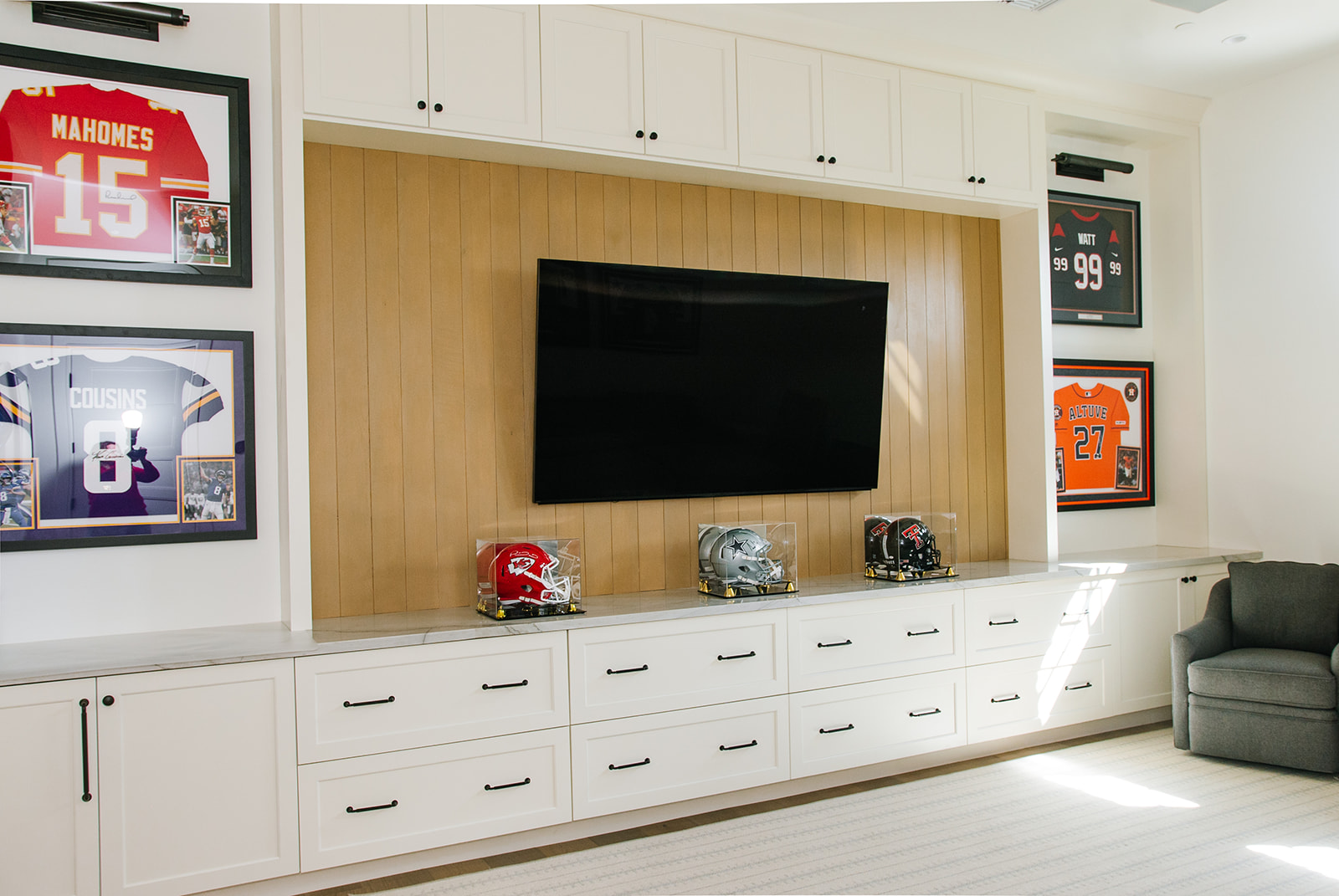 Sports-Themed Entertainment Room with Custom Cabinetry – A modern entertainment space featuring a large flat-screen TV, custom-built white cabinetry, and framed sports jerseys. Glass-encased football helmets on display add to the sports-themed decor.