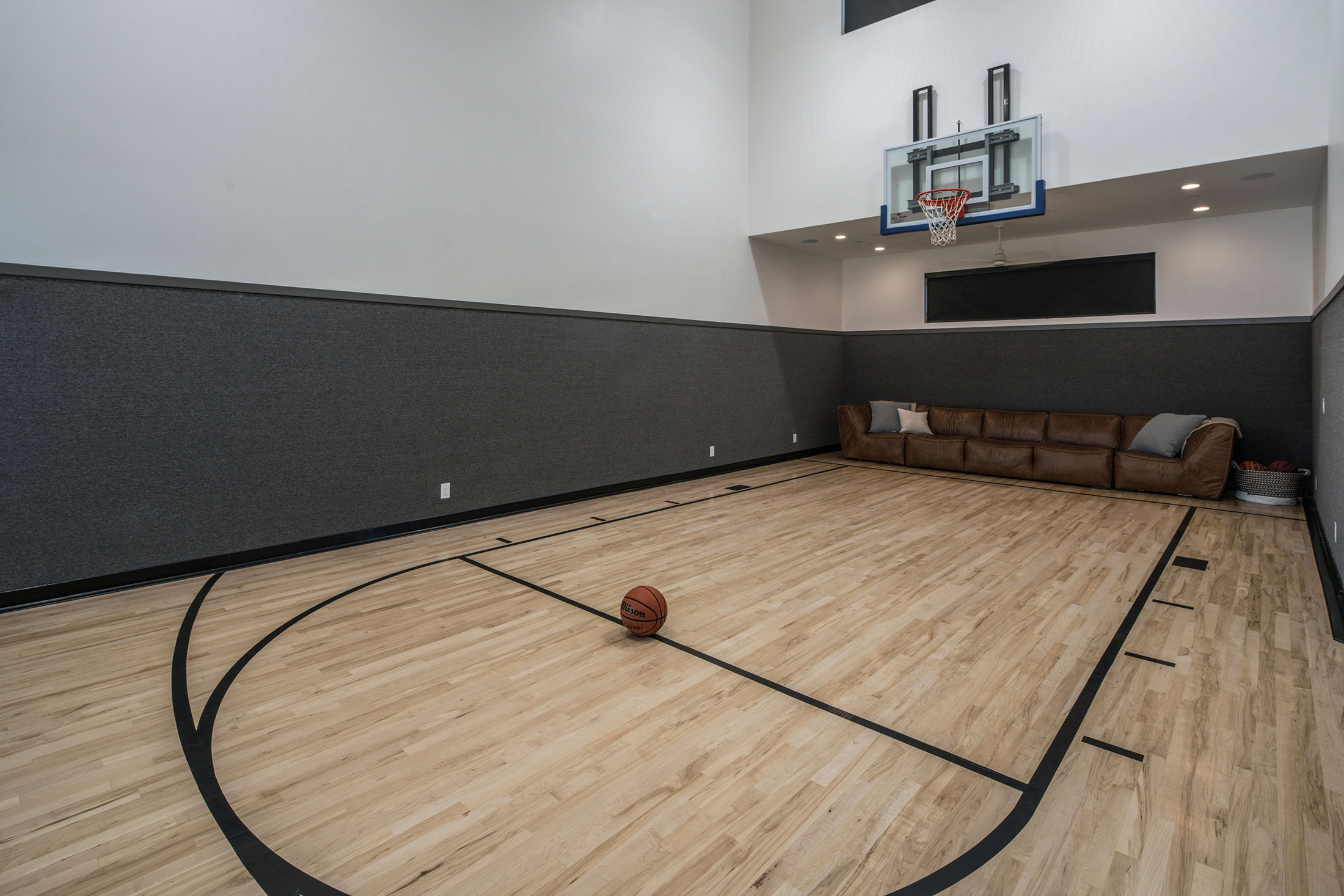 Indoor Basketball Court with Leather Couch and Hardwood Flooring – A spacious indoor basketball court with a sleek hardwood floor, a mounted hoop, and a cozy brown leather couch for relaxation. The walls feature dark acoustic paneling, enhancing the space’s modern and functional design.