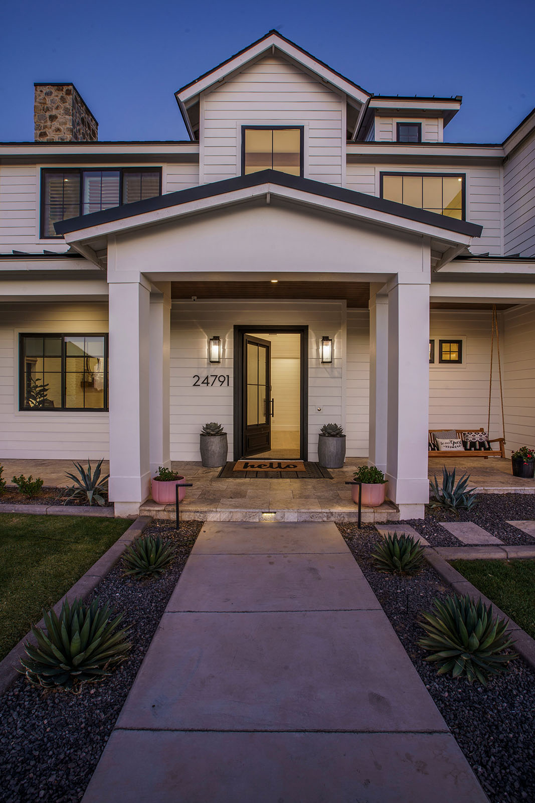 Elegant farmhouse entryway at dusk – A welcoming front entrance of a modern farmhouse featuring a covered porch, white siding, black trim, and a warmly lit doorway.