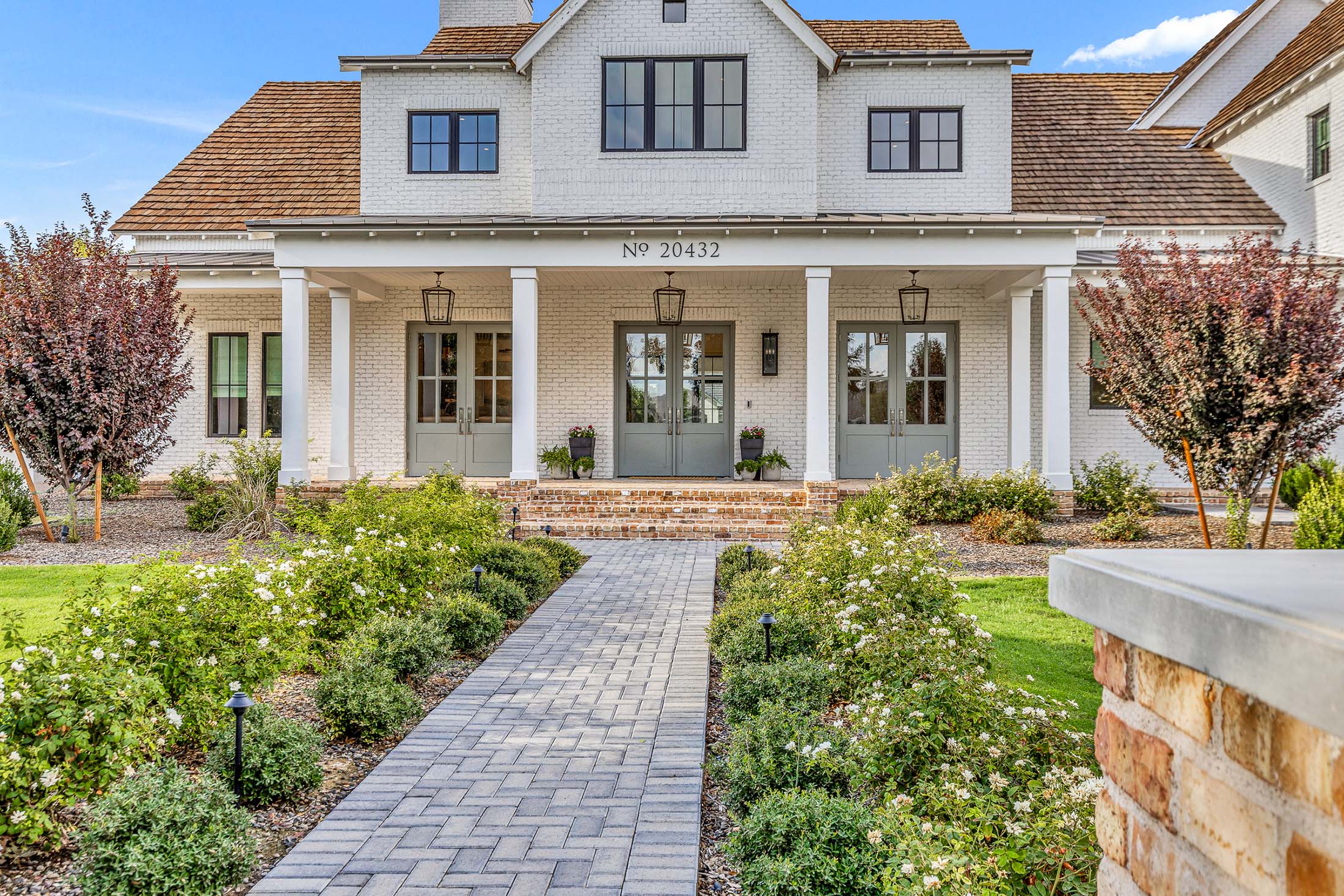 Luxury custom white brick home with a welcoming front porch, elegant landscaping, and timeless architectural details. Featuring classic wood accents, large glass-paneled doors, and lush greenery, this high-end residence embodies refined craftsmanship and sophisticated living.