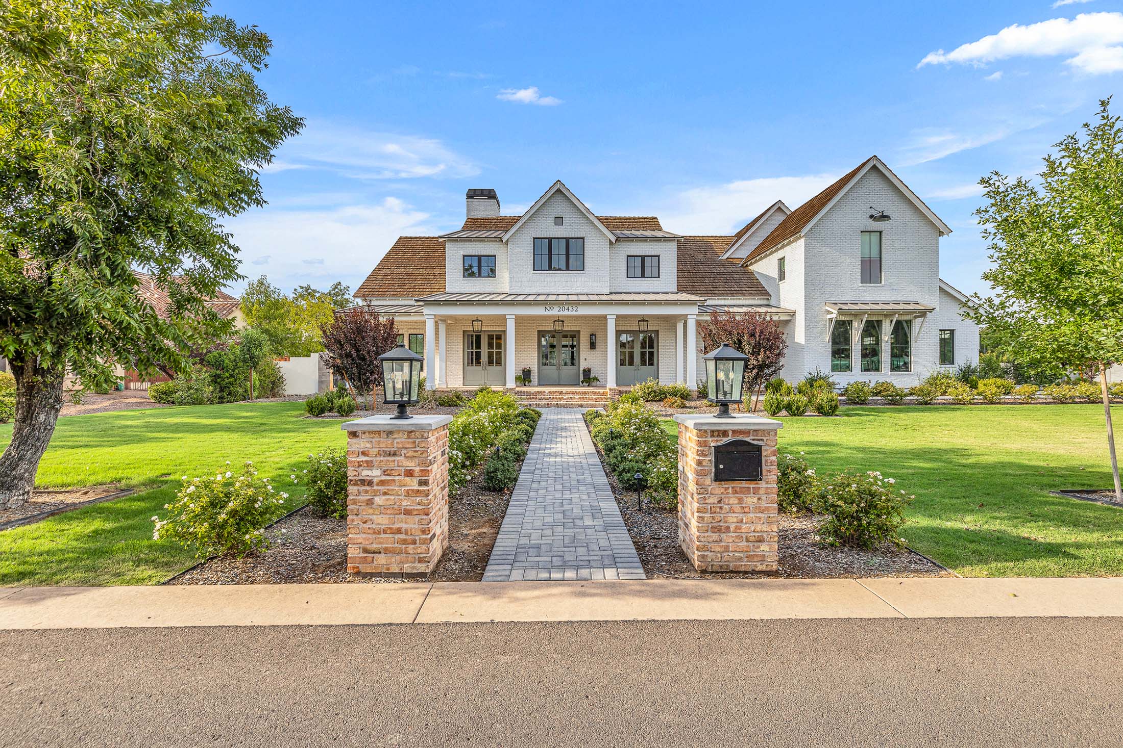 Luxury custom white brick home with a welcoming front porch, elegant landscaping, and timeless architectural details. Featuring classic wood accents, large glass-paneled doors, and lush greenery, this high-end residence embodies refined craftsmanship and sophisticated living.