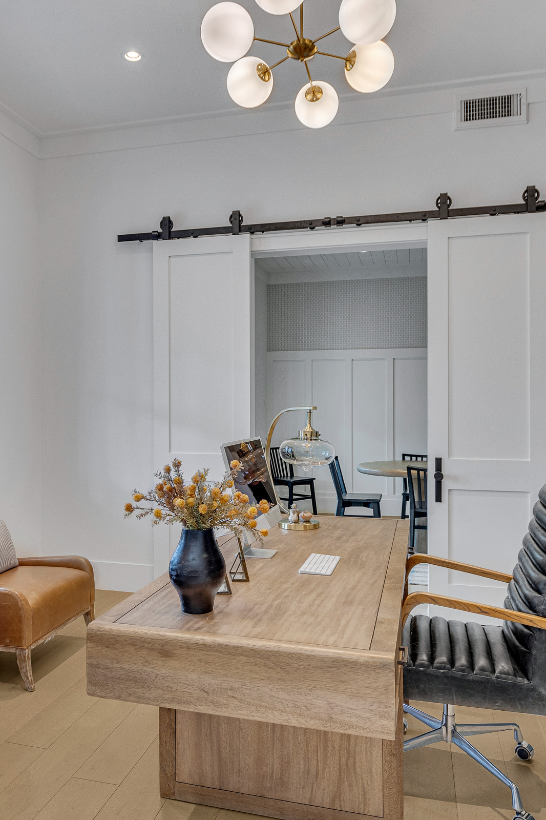 Sophisticated home office featuring a natural wood desk, built-in sage green cabinetry, and leather accent chairs. The space is illuminated by a modern brass chandelier and large glass-paneled double doors, while sliding barn doors lead to an adjoining room, blending functionality with timeless elegance.