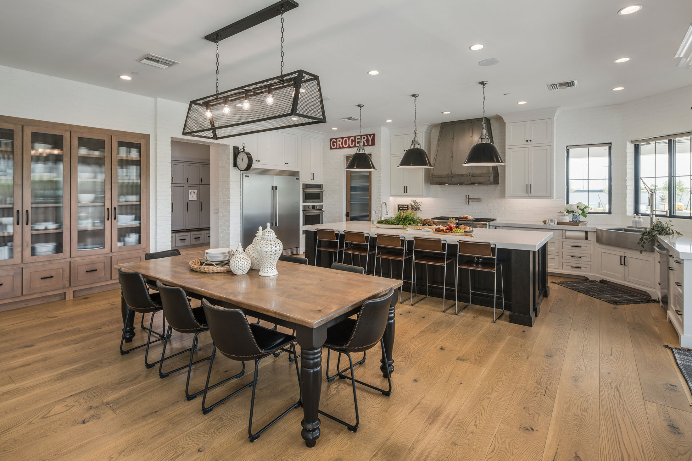 Elegant farmhouse dining and kitchen area with rustic wood elements – A warm and inviting dining space with a large wooden table, black chairs, and stylish industrial lighting. The open-concept kitchen boasts a white brick backsplash, a large range hood, and a walk-in pantry, blending farmhouse aesthetics with modern convenience.