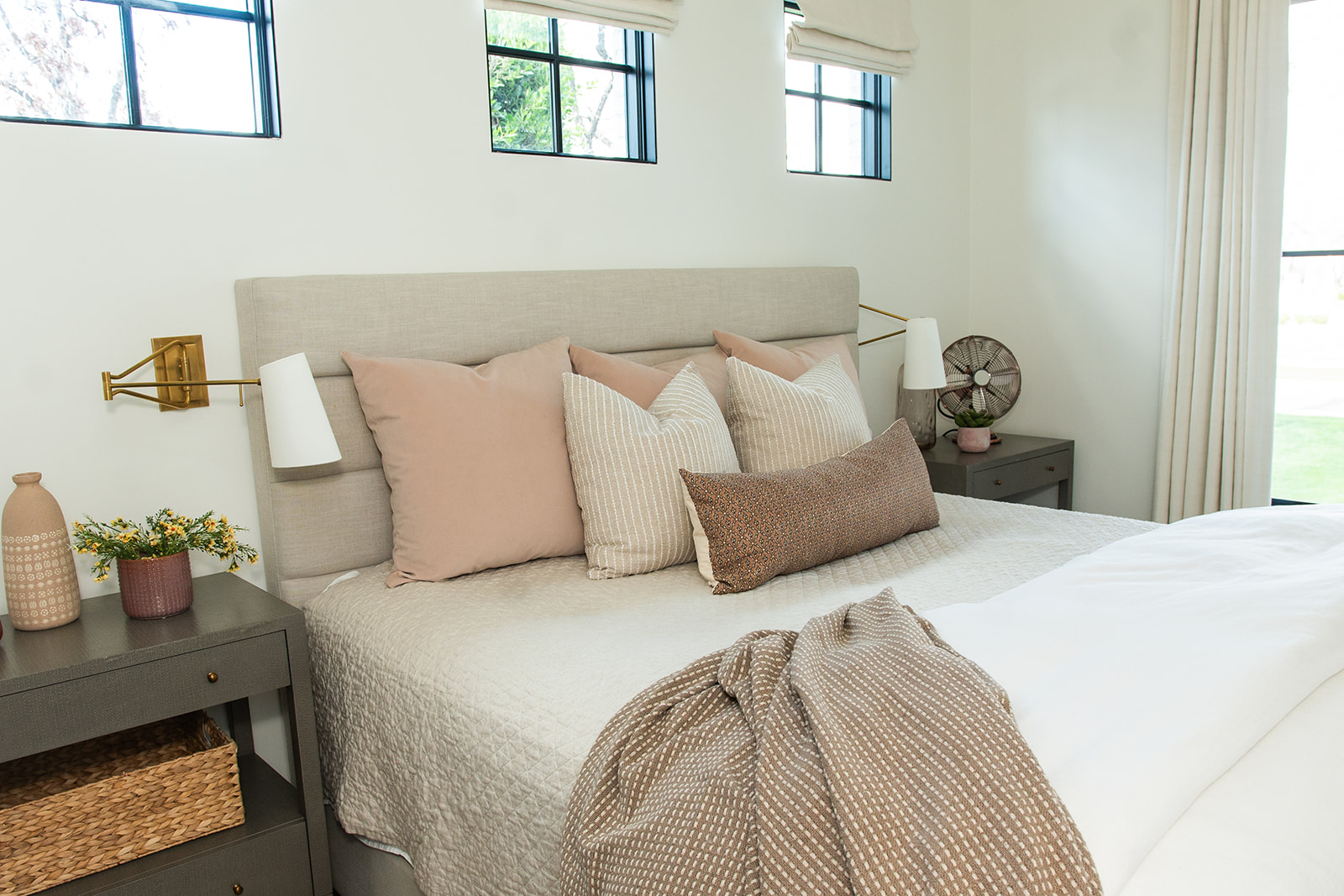 Cozy Neutral Bedroom with Soft Pink and Beige Accents – A beautifully styled bedroom featuring a light beige upholstered bed frame, soft pink and striped accent pillows, and brass swing-arm wall lamps. The modern bedside tables add a touch of elegance, complementing the minimalist decor.