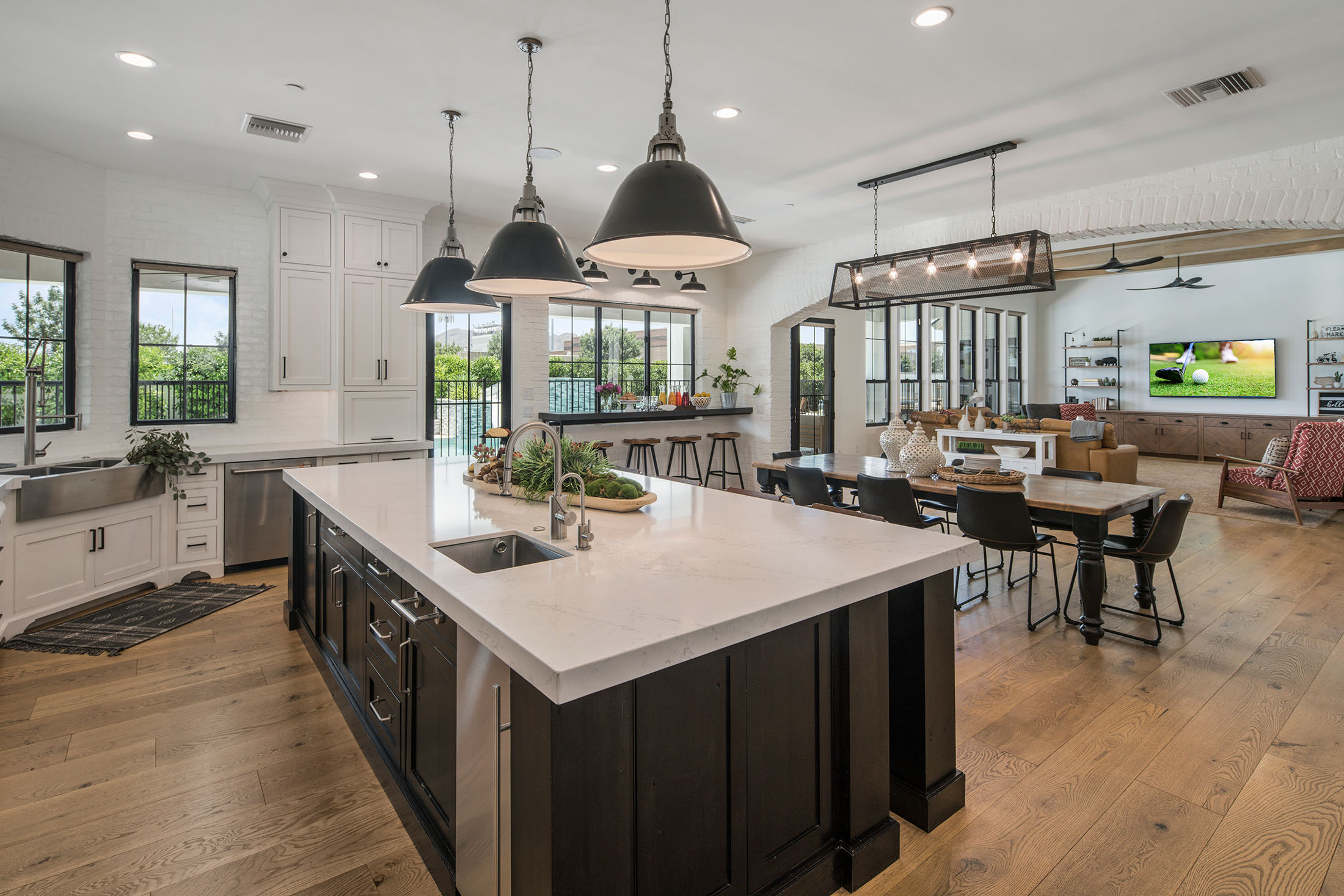 Spacious open-concept kitchen and living area featuring a large black and white island with quartz countertops, farmhouse sink, and industrial pendant lighting. The design blends modern and rustic elements with natural wood flooring, exposed brick, and sleek black-framed windows, creating a warm and inviting atmosphere perfect for entertaining.