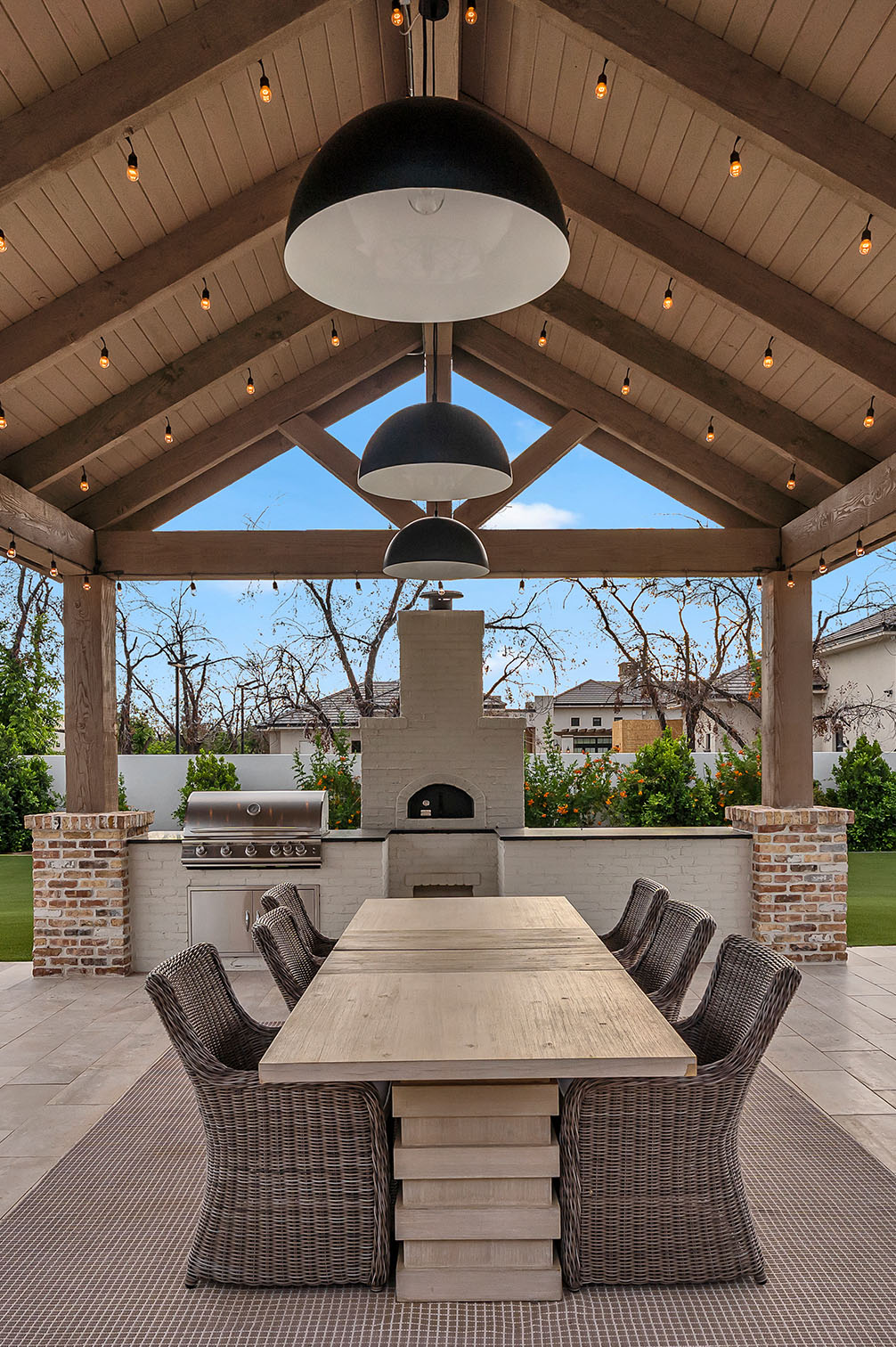 Outdoor dining area with a covered pergola, featuring a built-in stainless steel grill, white brick pizza oven, and a large wooden dining table with wicker chairs, illuminated by modern black pendant lights and string lights.