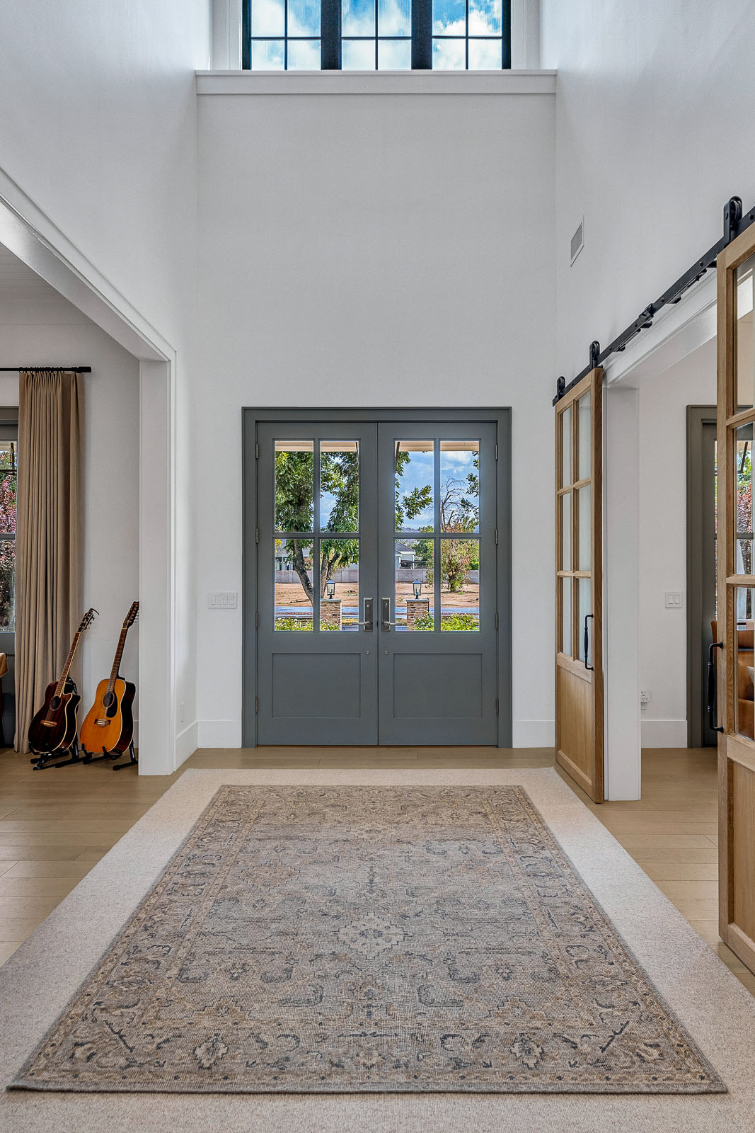 Grand entryway with double glass-paneled doors painted in muted sage green, high ceilings, and a large transom window allowing natural light to flood the space. Featuring a vintage-style area rug, warm wood flooring, and elegant sliding glass barn doors leading to adjacent rooms, this foyer blends timeless charm with modern sophistication.