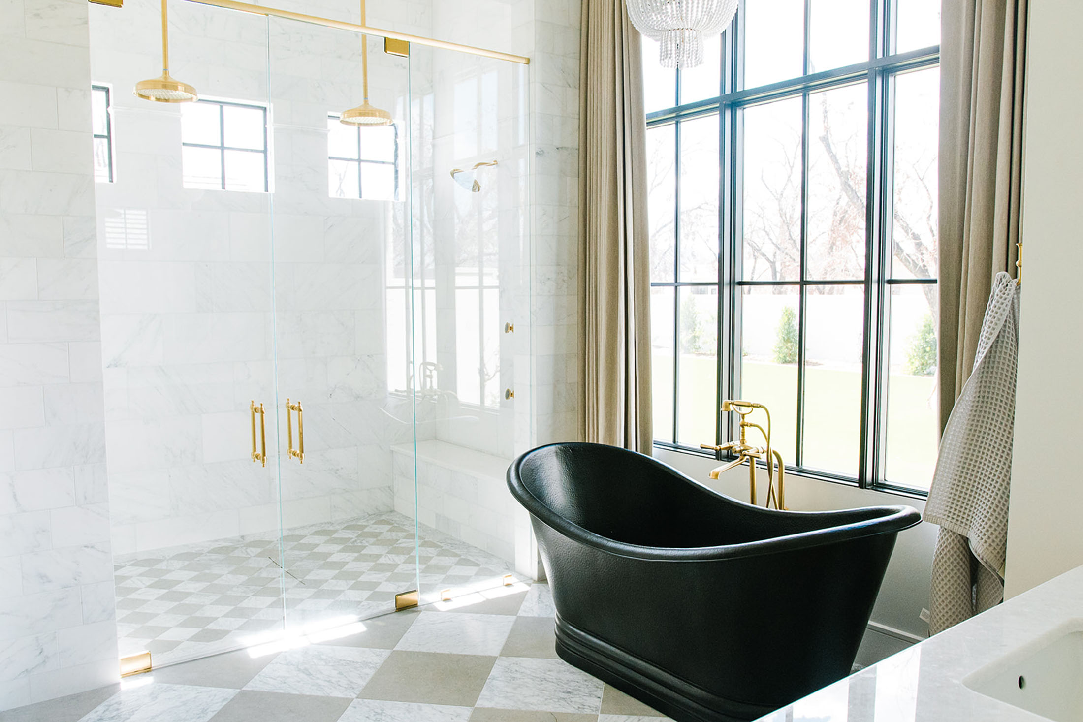 Elegant bathroom featuring a black freestanding soaking tub, a spacious glass-enclosed shower with gold fixtures, and floor-to-ceiling windows framed in black. The checkered marble flooring and crystal chandelier add a timeless, sophisticated touch.