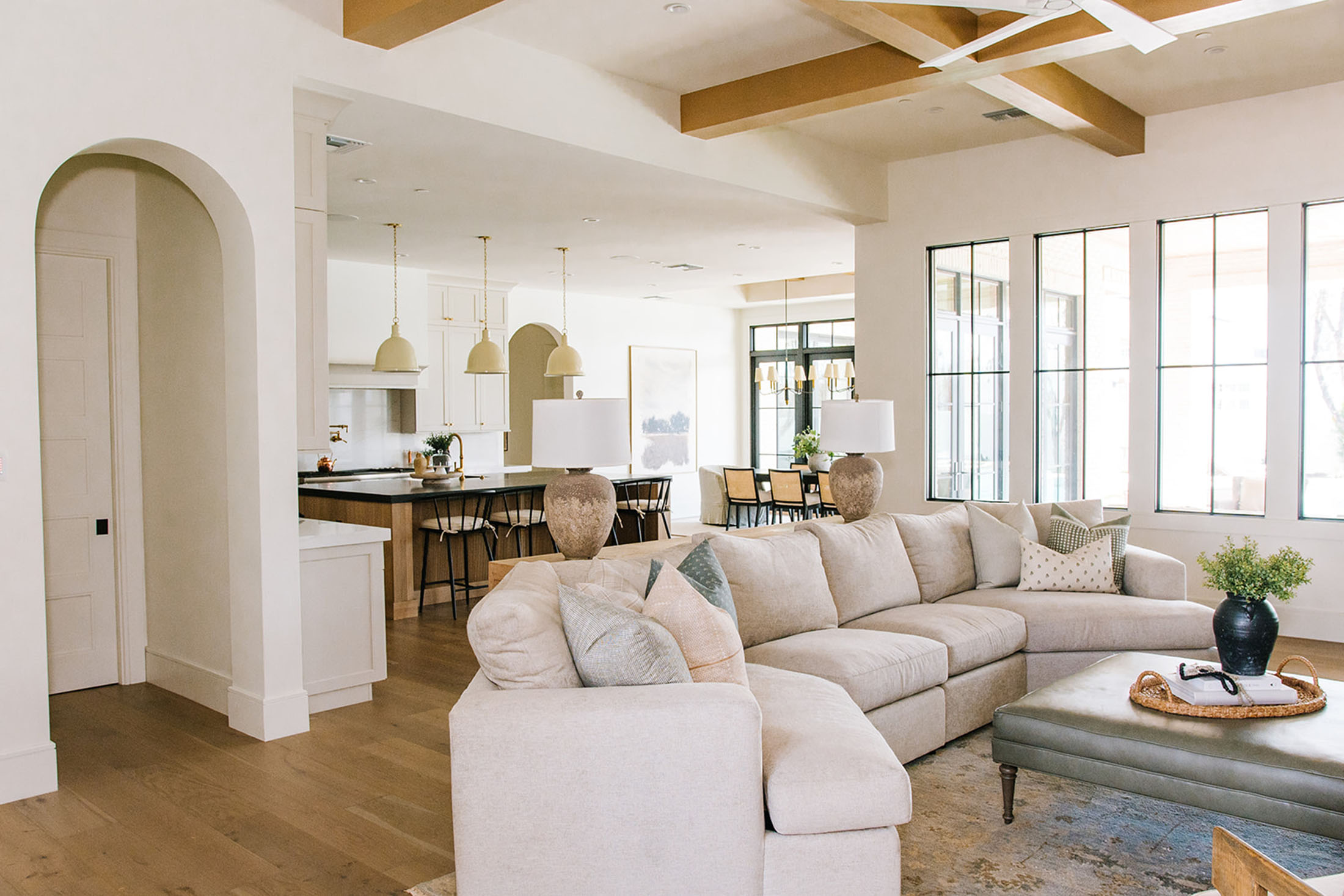 Spacious living area with a neutral sectional sofa, exposed wood ceiling beams, and large black-framed windows. The open floor plan seamlessly connects to the dining area and kitchen, creating a bright and inviting atmosphere.