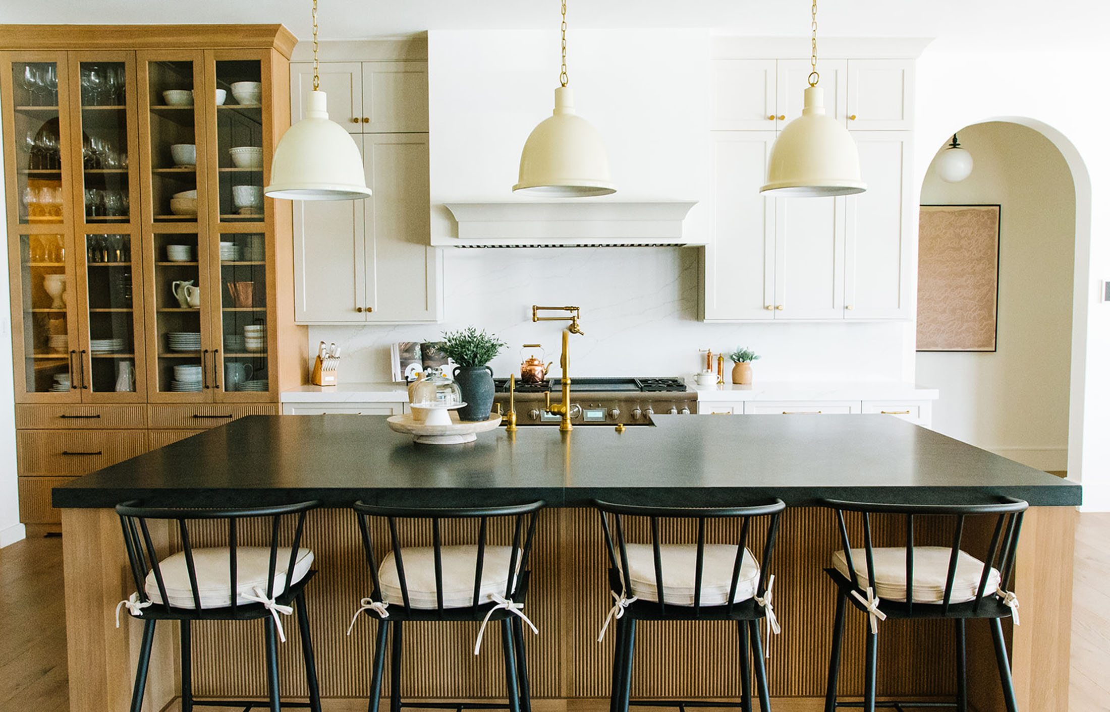 Stunning kitchen featuring a black quartz island, white shaker cabinets, and a built-in glass-front display for dinnerware. Soft gold pendant lights and brass hardware complement the warm wood tones, blending elegance with modern farmhouse charm.