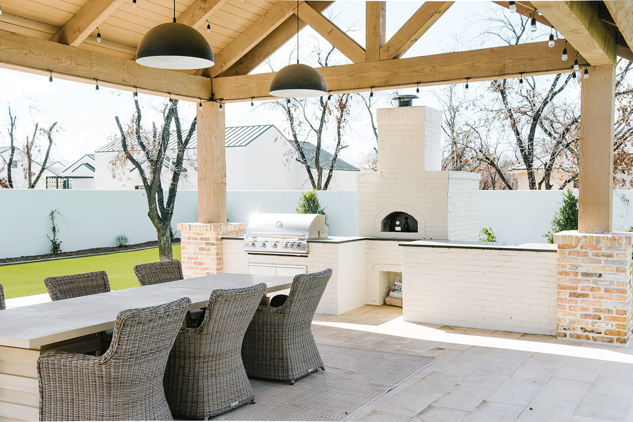 Expansive covered outdoor living space with a built-in brick pizza oven, stainless steel grill, and a rustic wood dining table surrounded by woven wicker chairs. The vaulted wood beam ceiling and black pendant lighting create a cozy yet refined entertaining space.