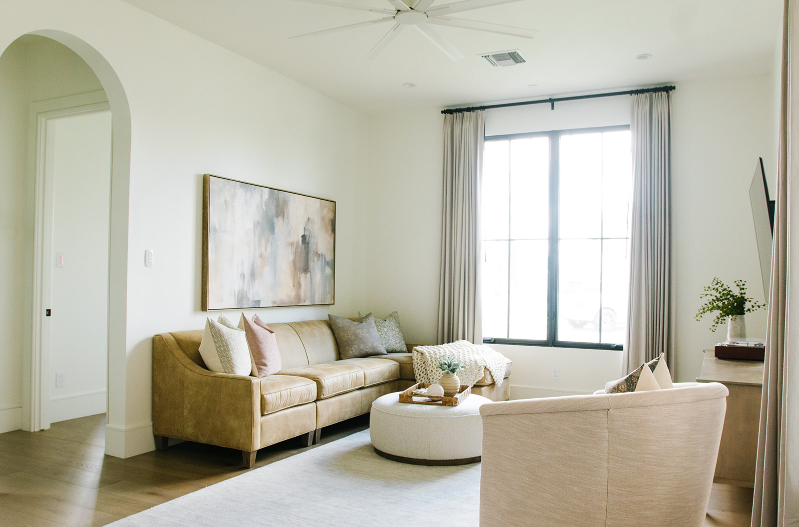 Bright and Elegant Living Room with Neutral Tones – A chic living space with a beige leather sofa, abstract wall art, and a round ottoman coffee table. Large windows with neutral drapes allow natural light to flood the room, enhancing its inviting ambiance.