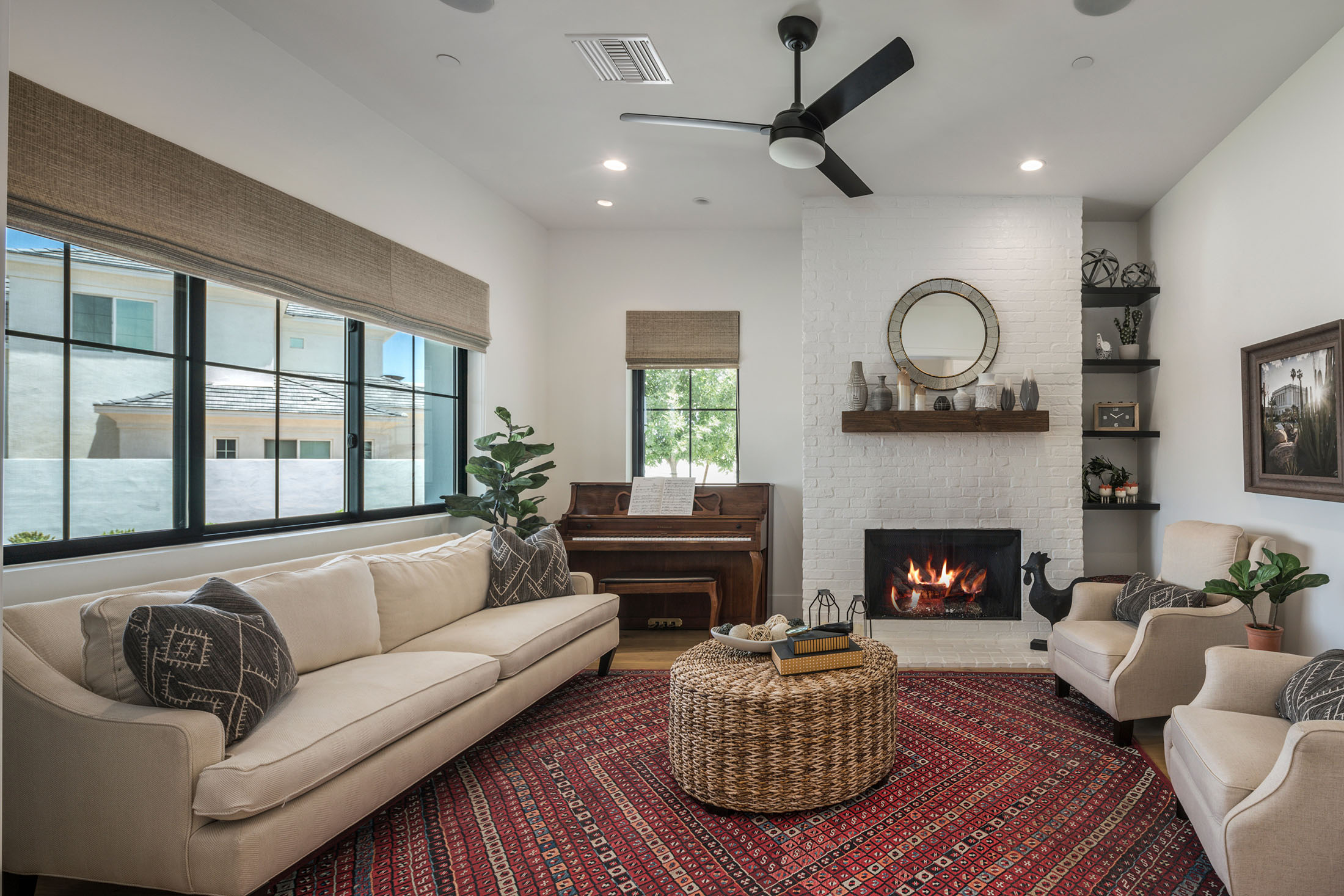 Cozy Living Room with Fireplace and Piano – A warm and inviting living room with a white brick fireplace, wooden mantel, and built-in shelving. A cream-colored sofa and armchairs surround a woven center table, while a grand piano adds a classic touch.