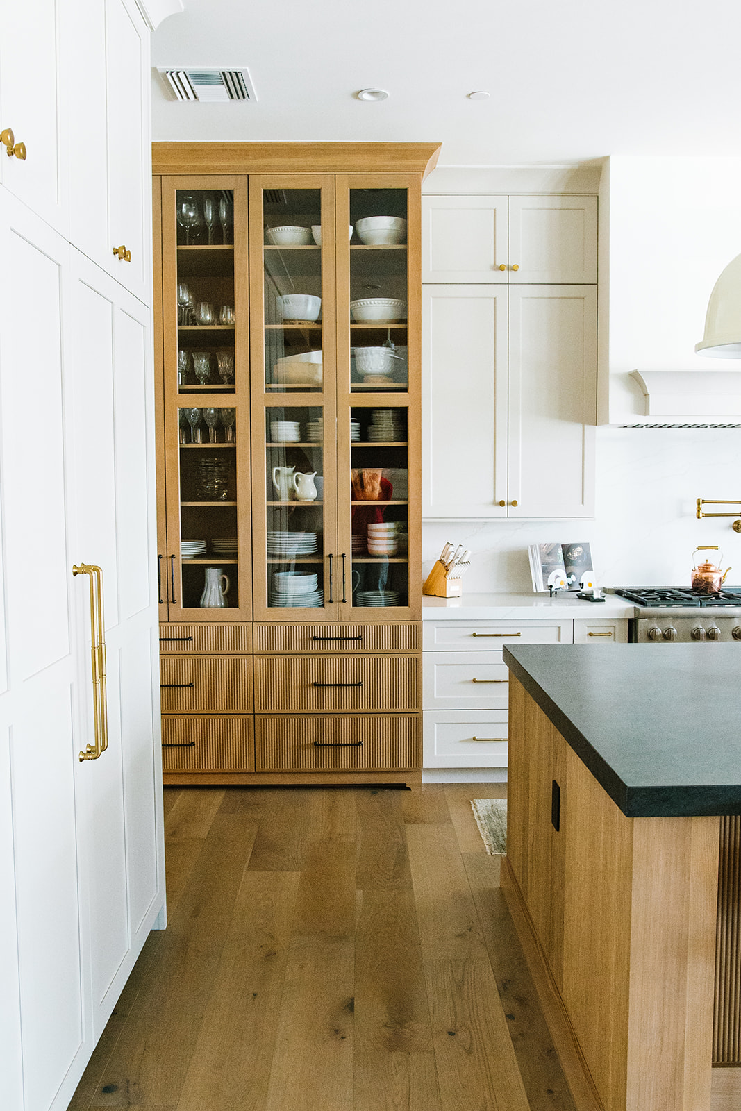 Stylish Kitchen with Glass Cabinetry and Natural Wood Accents – A beautifully designed kitchen featuring glass-front wooden cabinetry, white shaker cabinets, and a black stone countertop. Gold hardware and a white range hood enhance the bright, airy aesthetic.