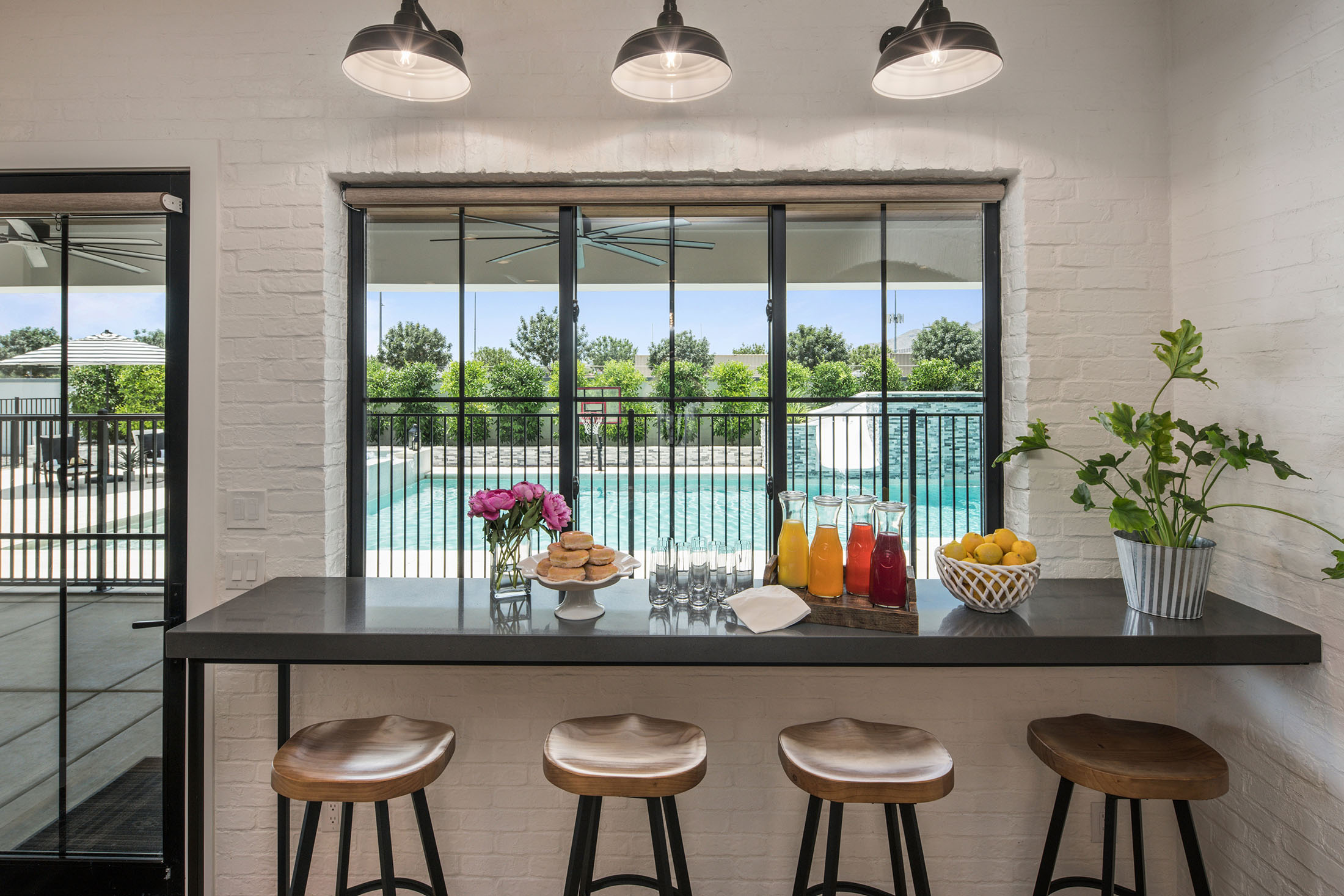 Poolside Breakfast Bar with Scenic Outdoor View – A charming indoor breakfast bar overlooking a luxurious backyard pool. A black countertop, wooden barstools, and a spread of fresh juice and pastries create a welcoming ambiance under industrial pendant lights.