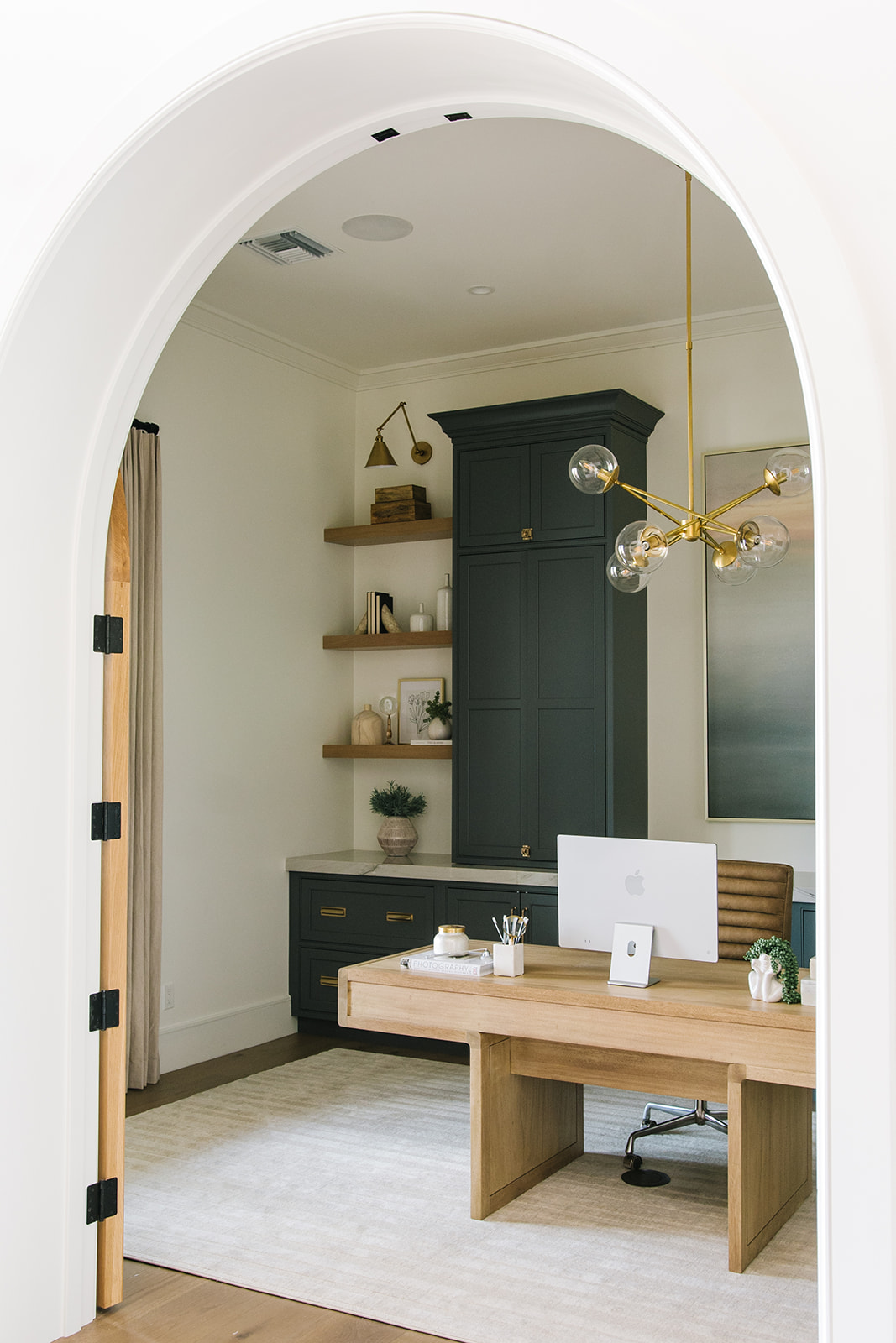 Sophisticated Home Office with Arched Doorway - A stylish home office with a natural wood desk, plush white armchairs, dark cabinetry, and floating shelves, framed by a striking arched entryway.