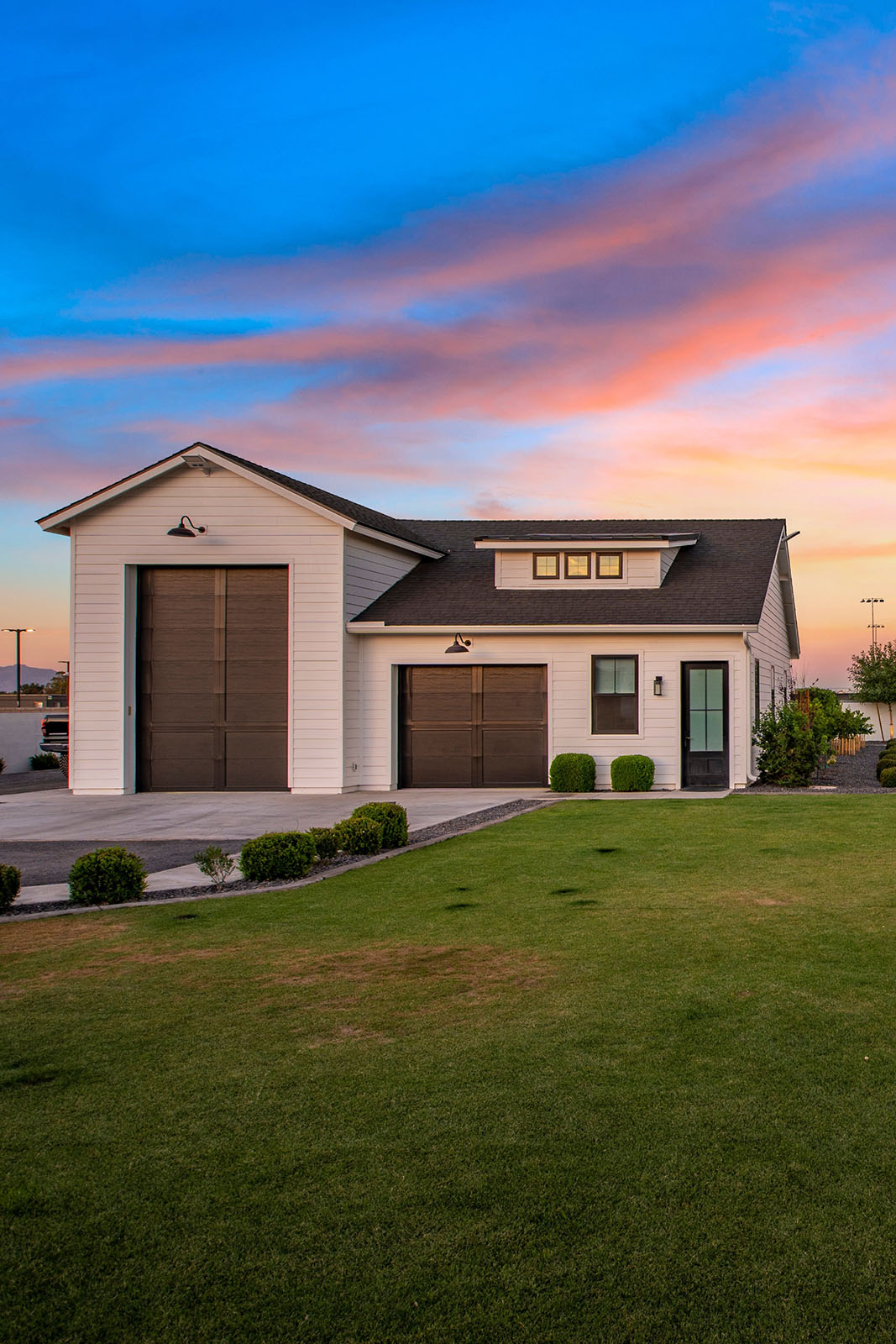 Modern farmhouse-style garage at sunset – A white detached garage with large brown doors, black exterior lights, and a well-manicured lawn, set against a vibrant pink and blue sunset sky.