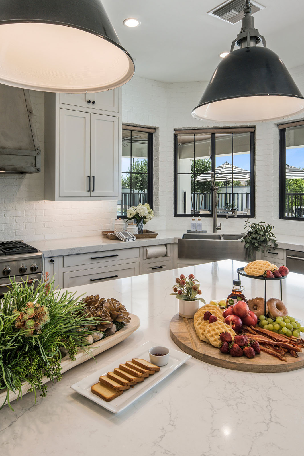 Modern farmhouse kitchen with a large white marble island and breakfast spread – A beautifully designed kitchen featuring a spacious marble island, industrial black pendant lighting, and an inviting breakfast spread with fresh fruits, waffles, and bagels. Large windows with black trim bring in natural light, complementing the white cabinetry and farmhouse sink.