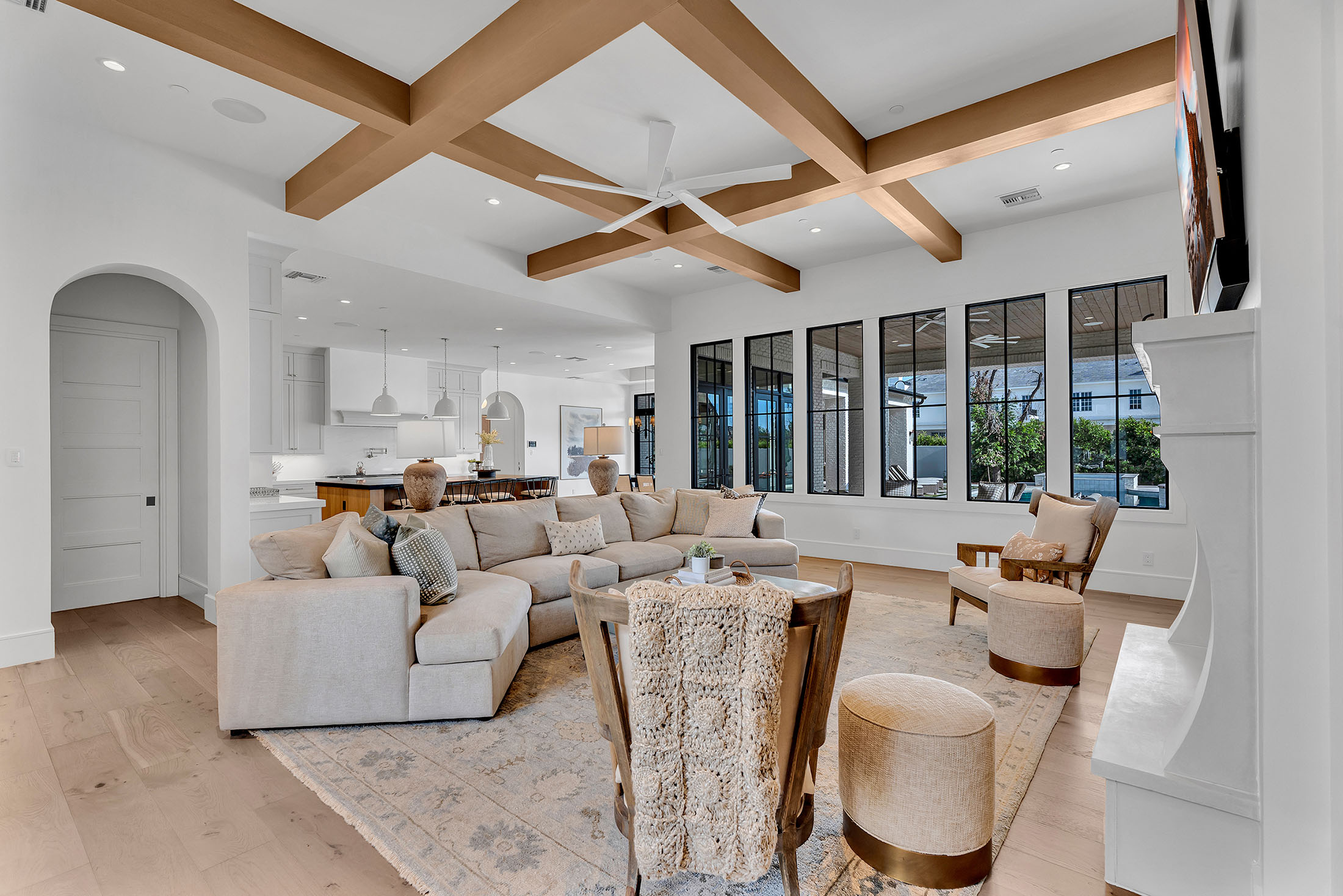 Spacious living area with a neutral sectional sofa, exposed wood ceiling beams, and large black-framed windows. The open floor plan seamlessly connects to the dining area and kitchen, creating a bright and inviting atmosphere.