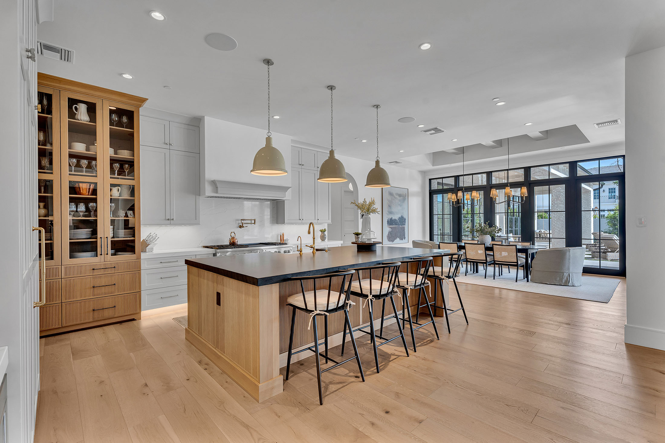 Stunning kitchen featuring a black quartz island, white shaker cabinets, and a built-in glass-front display for dinnerware. Soft gold pendant lights and brass hardware complement the warm wood tones, blending elegance with modern farmhouse charm.