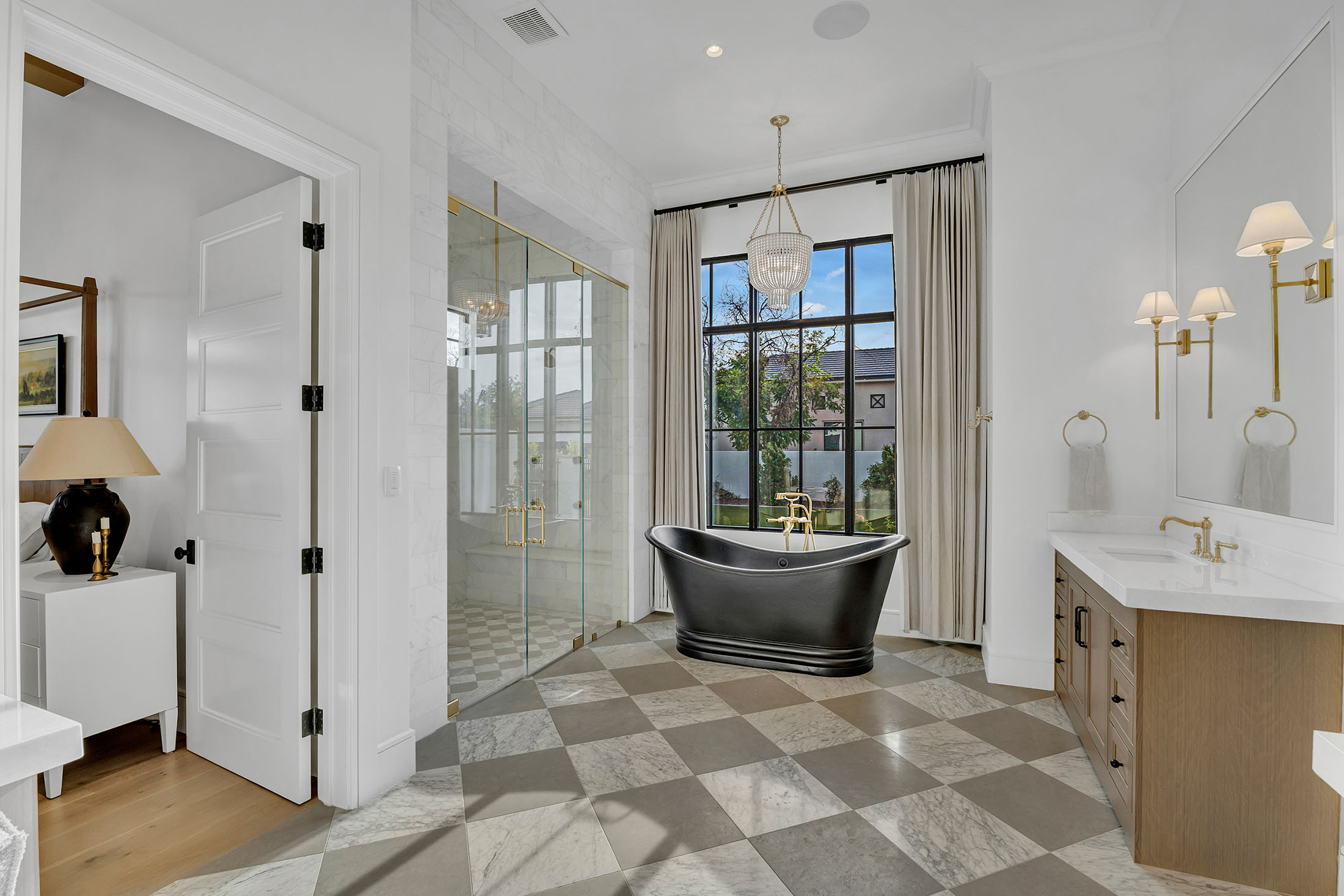 Elegant bathroom featuring a black freestanding soaking tub, a spacious glass-enclosed shower with gold fixtures, and floor-to-ceiling windows framed in black. The checkered marble flooring and crystal chandelier add a timeless, sophisticated touch.