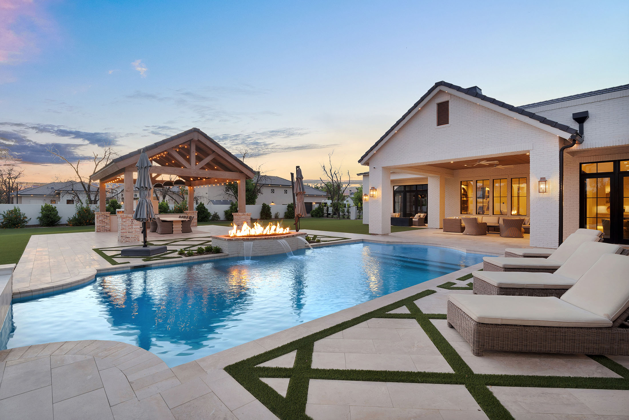 Twilight view of a luxury Arizona backyard oasis featuring a modern swimming pool with integrated fire and water features, custom travertine pavers with turf inlays, and a covered outdoor kitchen pavilion with string lighting. Includes a white brick contemporary home with expansive windows, a shaded patio lounge, and upscale poolside furniture — showcasing premier outdoor living design by The Ashtin Group.